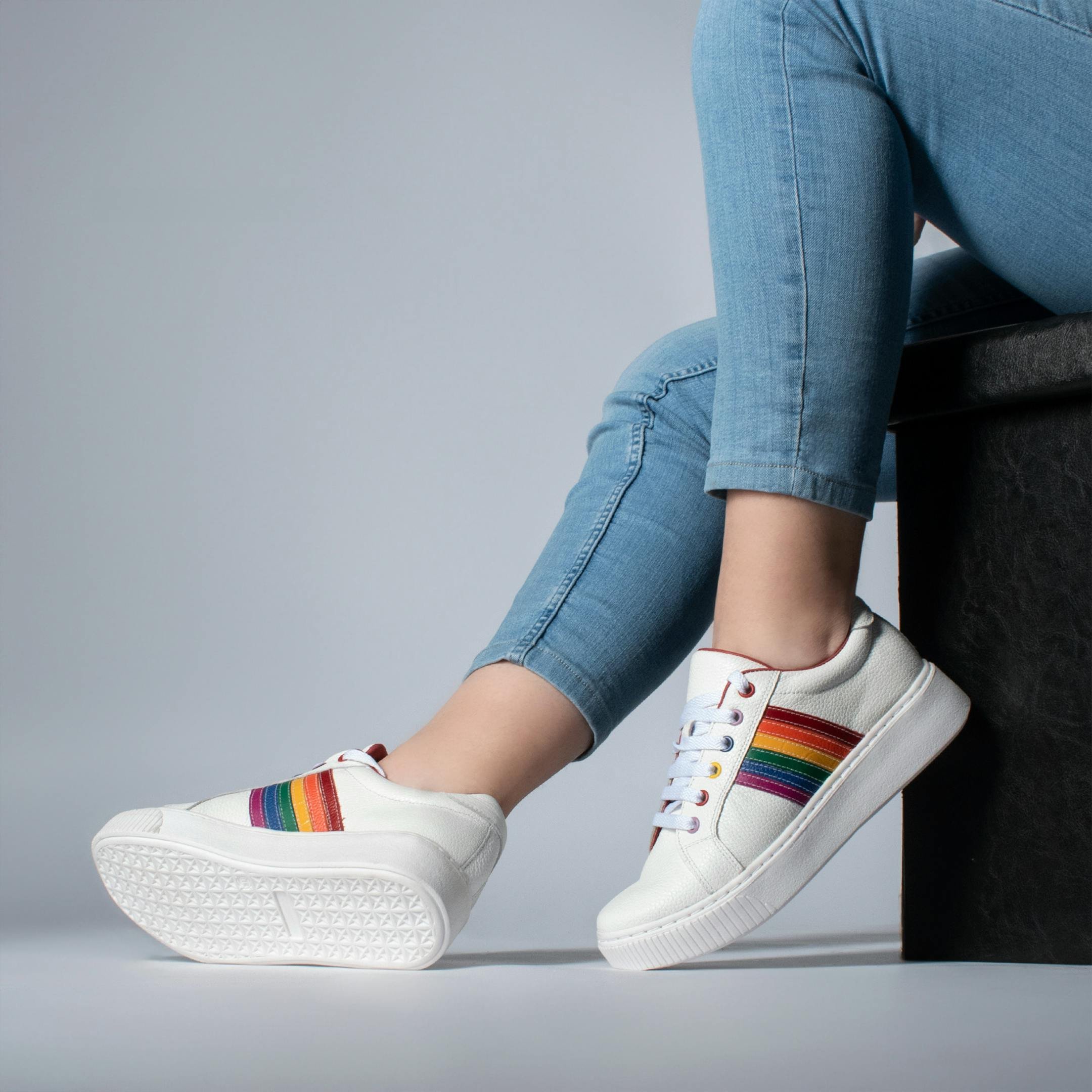 Close-up of white sneakers with rainbow-colored stripes and light blue denim jeans, against a gray background.
