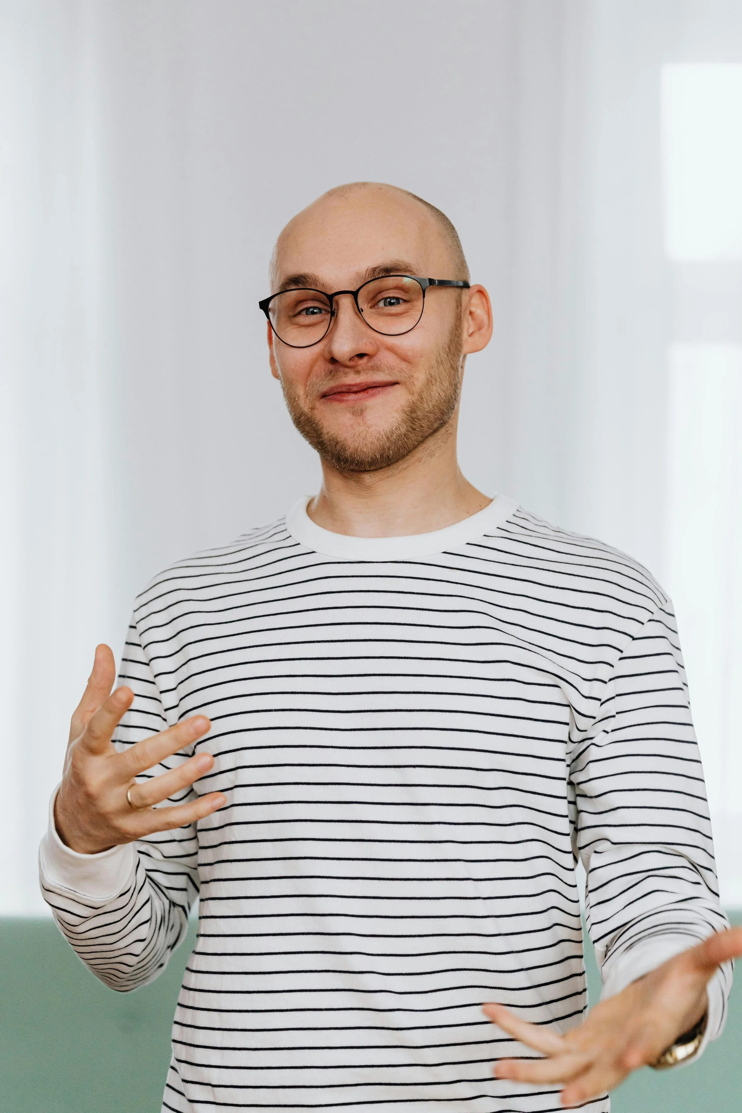 A young man with glasses, a beard, and a shaved head, smiling and making a peace sign with his fingers, wearing a white shirt with black horizontal stripes, standing indoors.