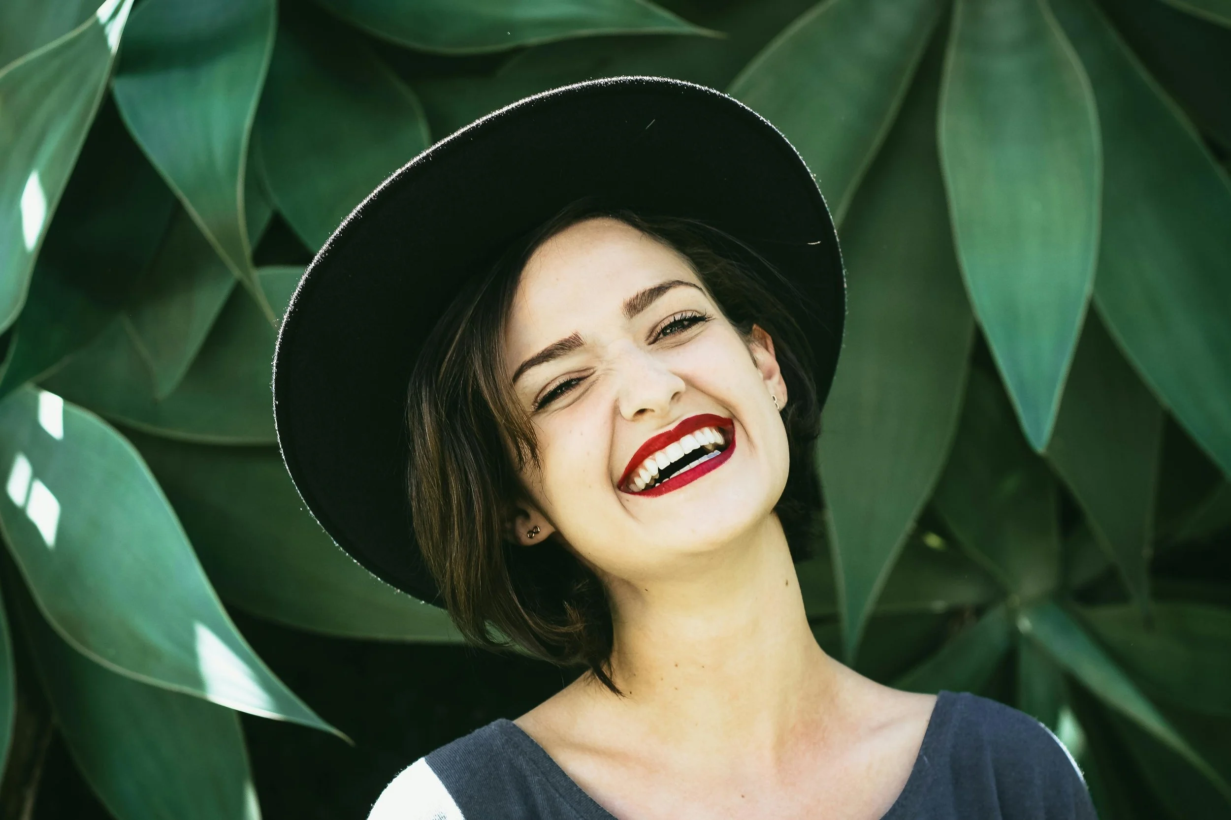A woman with short dark hair, wearing a black wide-brimmed hat, smiling widely with red lipstick, standing in front of large green leaves.