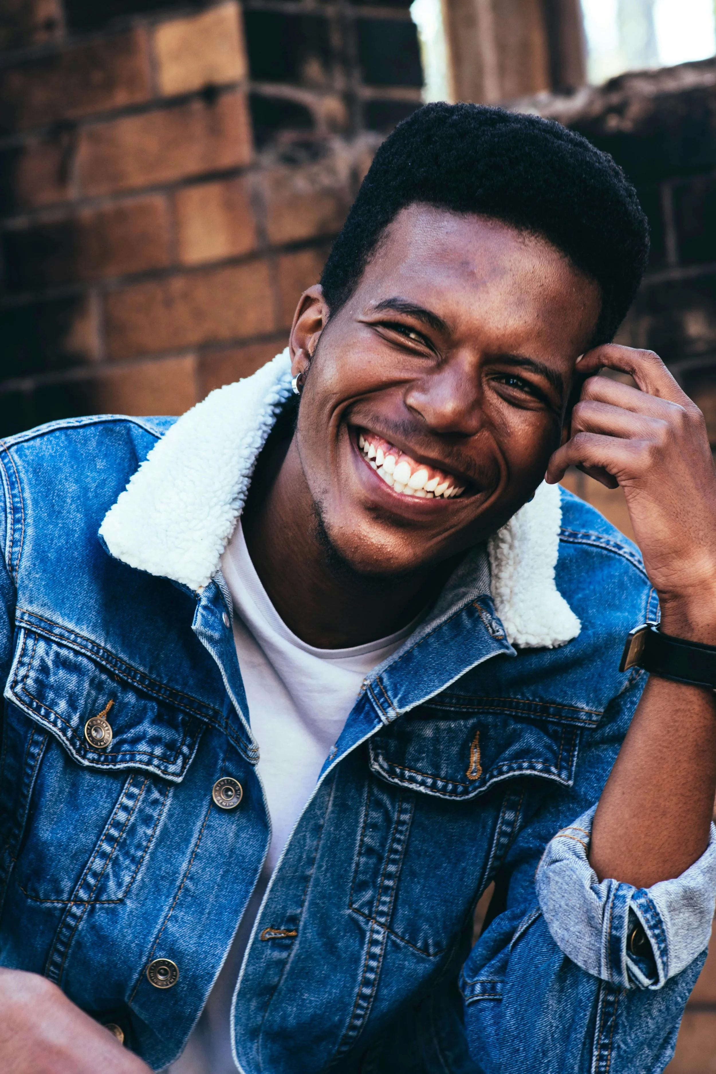 A young person with short, black hair smiling broadly, showing teeth, wearing a denim jacket with a white wool collar, seated outdoors near a brick wall.