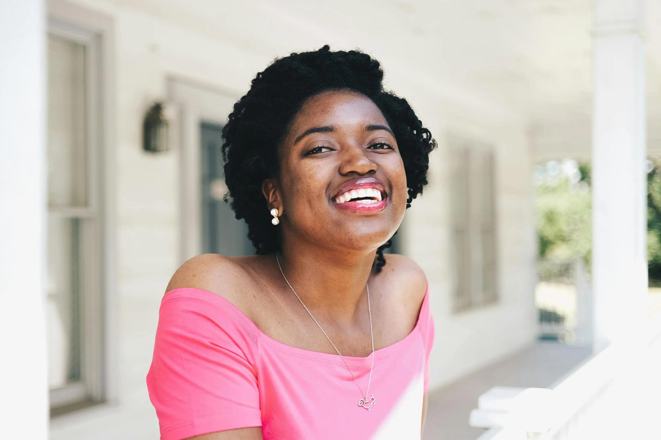 A woman with dark curly hair, wearing a pink top, earrings, and a necklace, standing outside near a white building with columns, smiling with teeth showing.