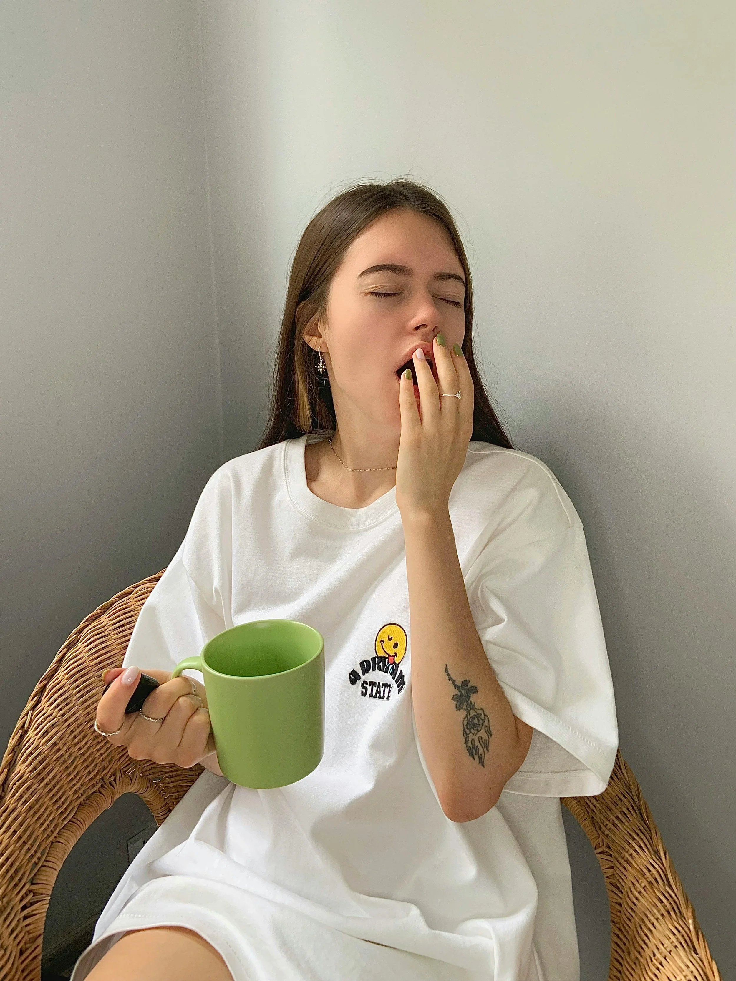 Young woman with tattoos and earrings sitting in a wicker chair, holding a green mug, yawning with eyes closed, wearing a white t-shirt with a smiley face and text.