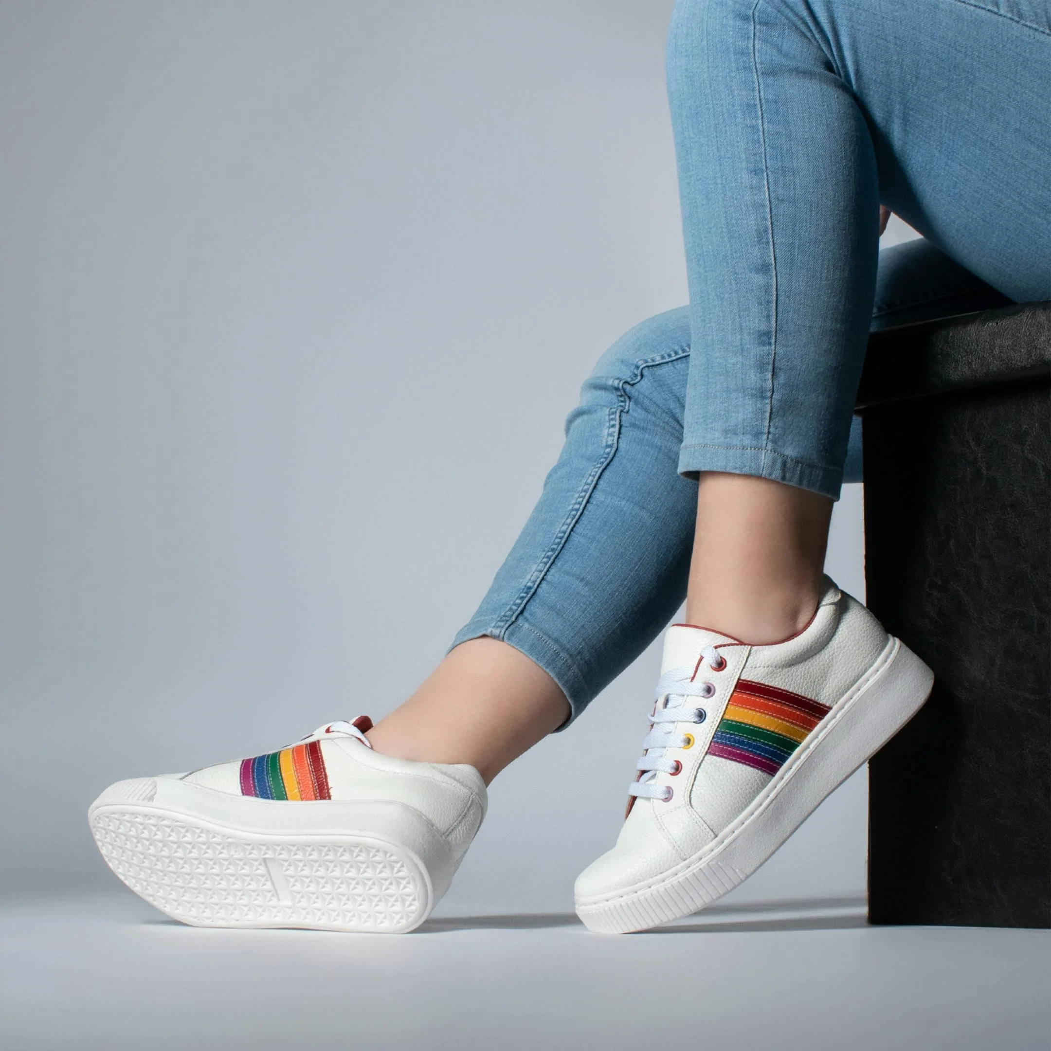 A person sitting on a black stool wearing electric blue jeans and white sneakers with rainbow-colored stripes.
