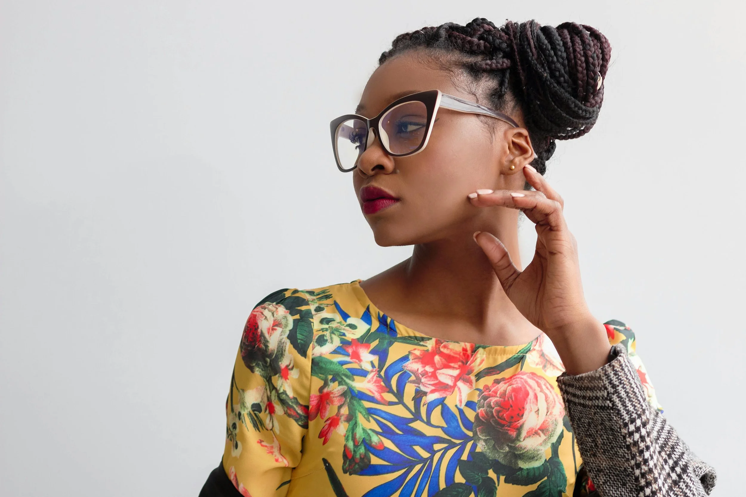 A woman with braided hair styled in an updo, wearing large sunglasses and a floral-patterned yellow top, touching her ear with her right hand, against a plain white background.