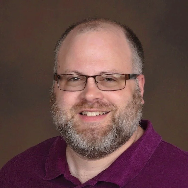 A man with glasses and a beard smiling, wearing a purple collared shirt.