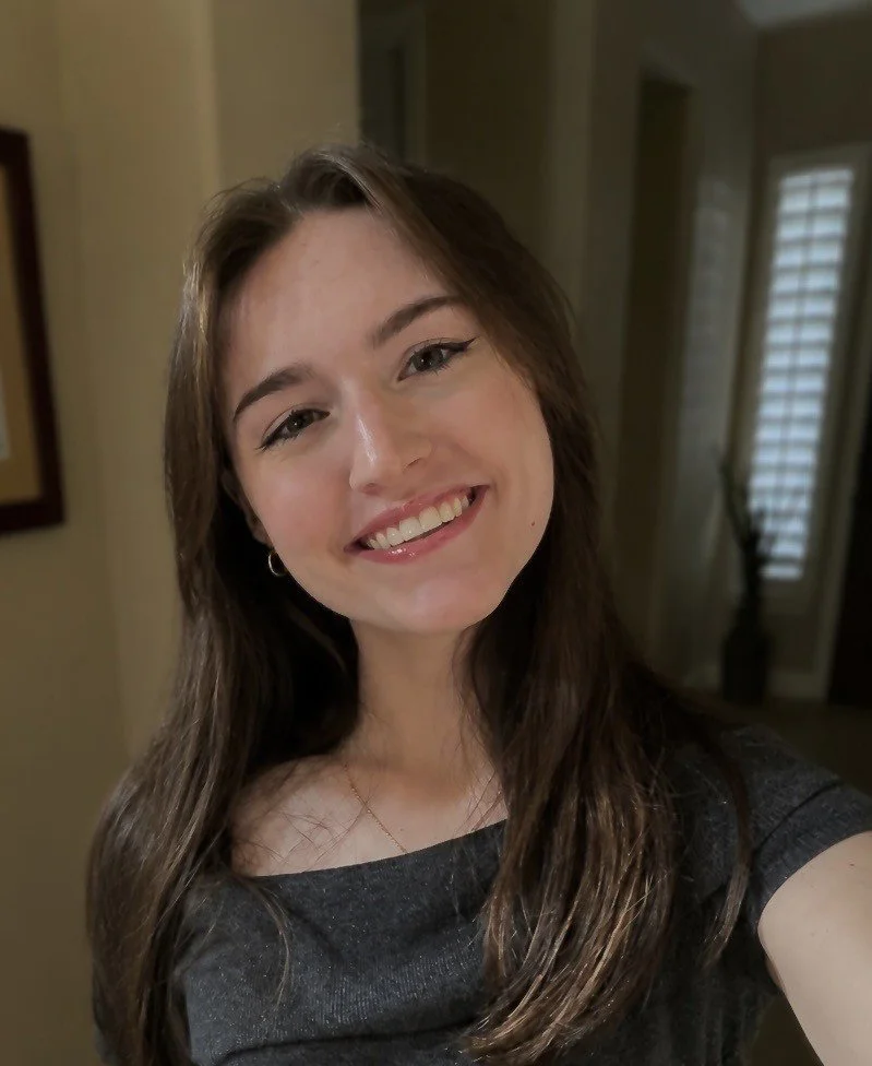 Kendal Black, a smiling young woman with long brown hair, wearing a dark top, standing indoors near windows with blinds