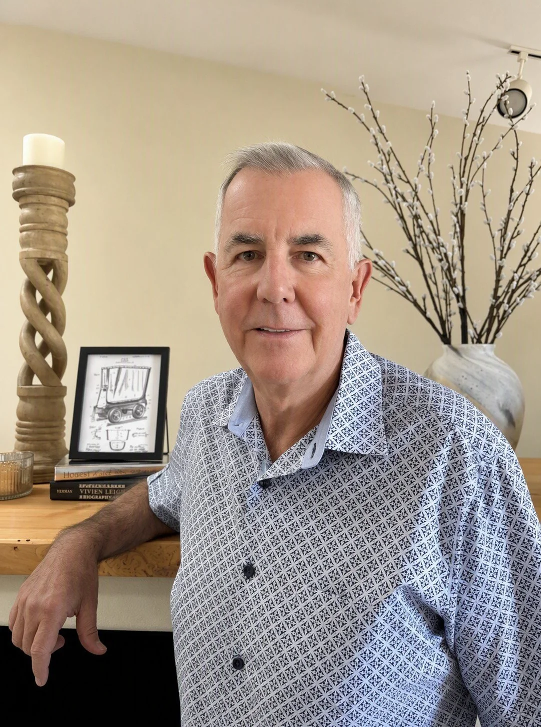 Patrick Findlay, a middle-aged man with gray hair in a patterned button-up shirt sits at a wooden table, smiling at the camera, with decorative branches in a white vase and picture frames in the background.