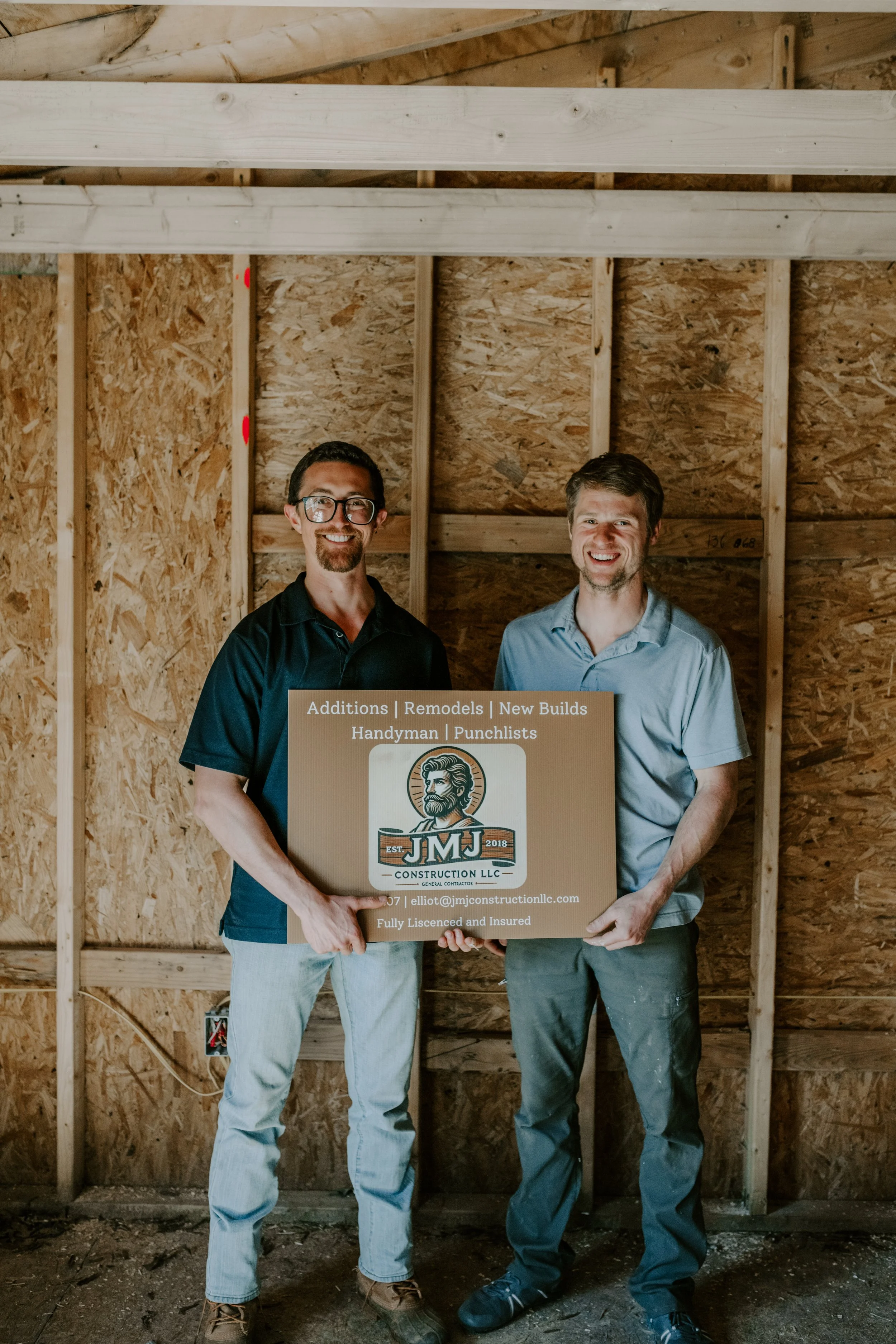 Two men smiling and holding a sign in a construction site with unfinished wooden walls. The sign reads 'Additions | Remodels | New Builds | Handyman | Punchlists' and features a logo of a bearded man with the text 'J.M.J Construction LLC'