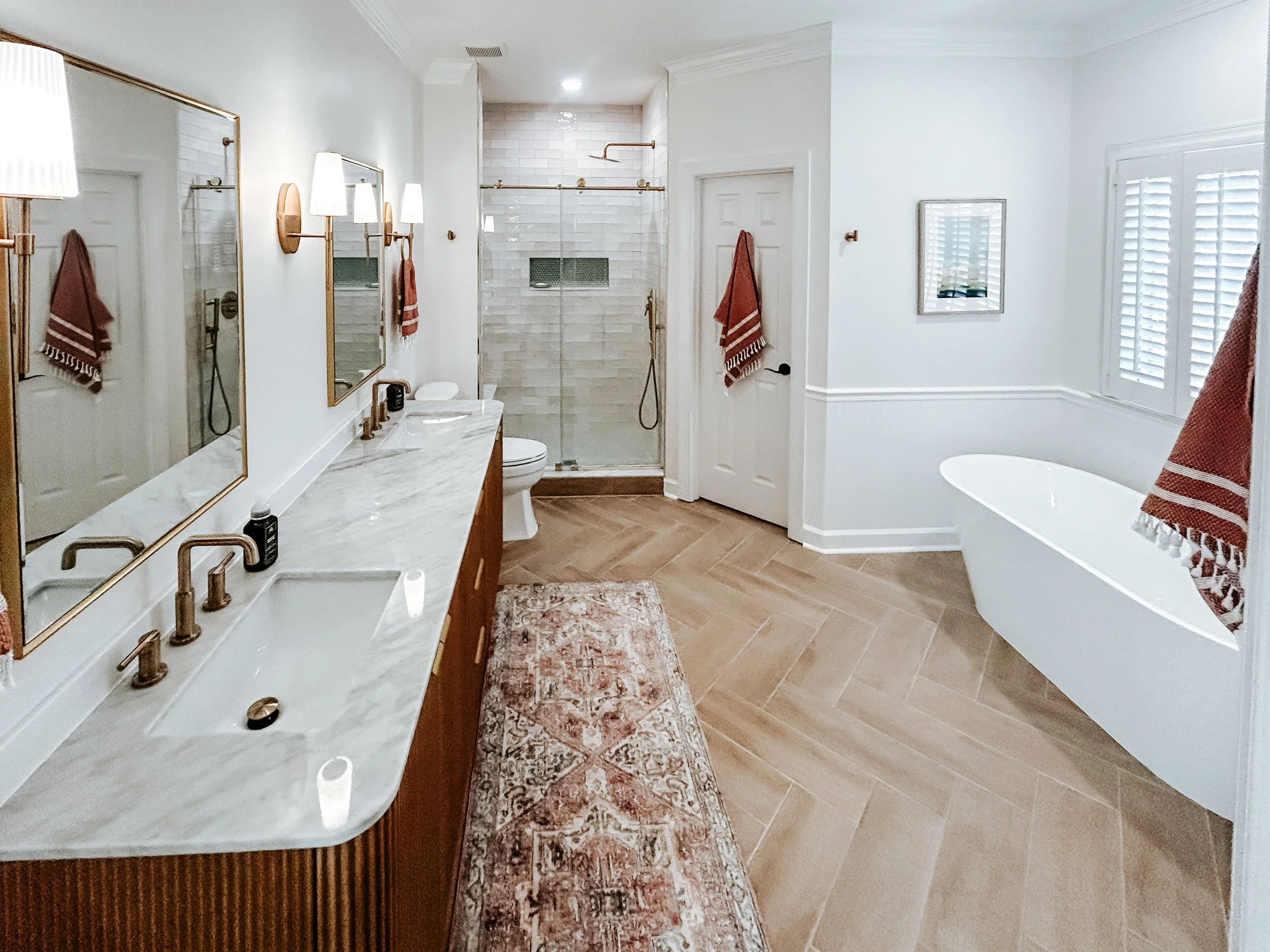 Bright, spacious bathroom featuring a double vanity with marble countertop, gold fixtures, two mirrors, and wall-mounted lights. There is a freestanding bathtub next to a window with plantation shutters. A glass-enclosed shower with a built-in niche is in the background. The floor is wood-look tile, and there is a patterned rug in the center.