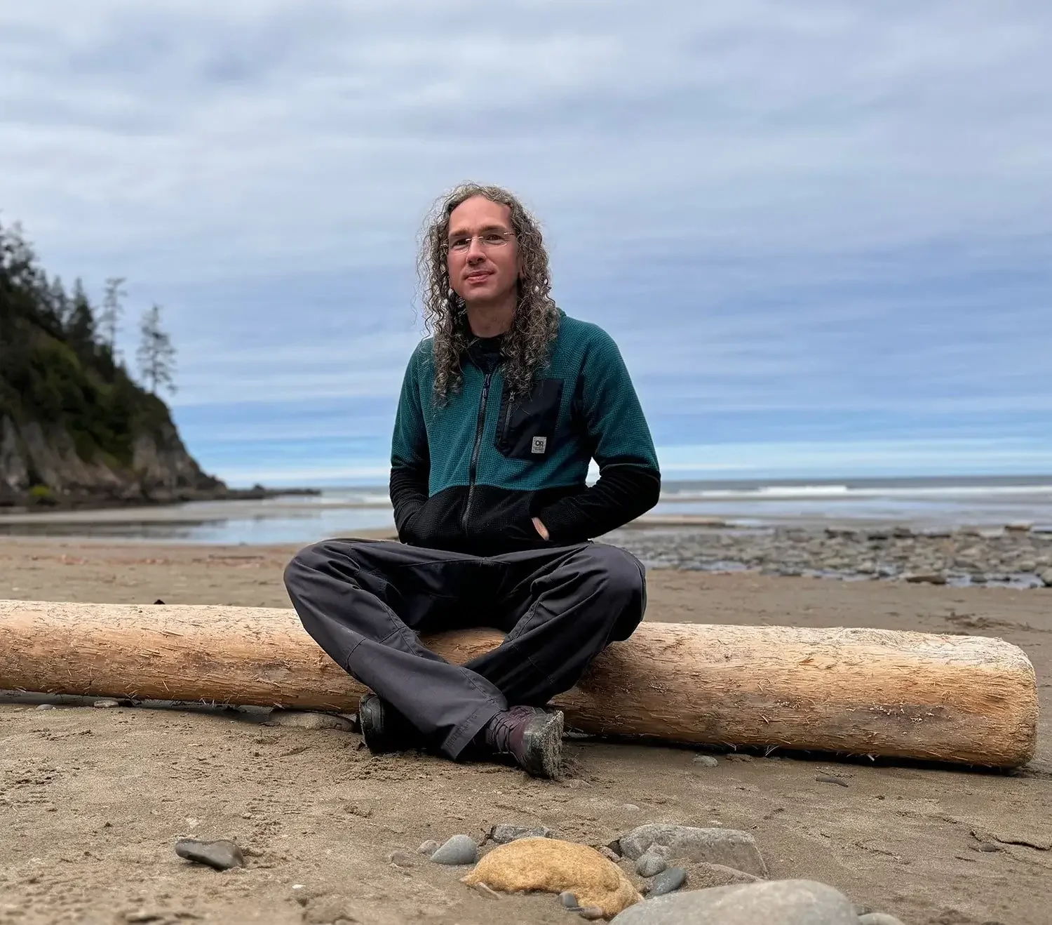 Jesse sits cross-legged on a log on a beach, with the ocean and a cloudy sky in the background.
