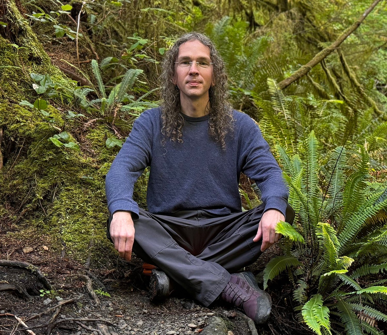 Jesse Deal sits cross-legged on the ground in a wooded area surrounded by ferns and trees. The individual has long curly hair, wears glasses, a blue sweater, and dark pants. The background is lush with greenery, including moss and various plants.