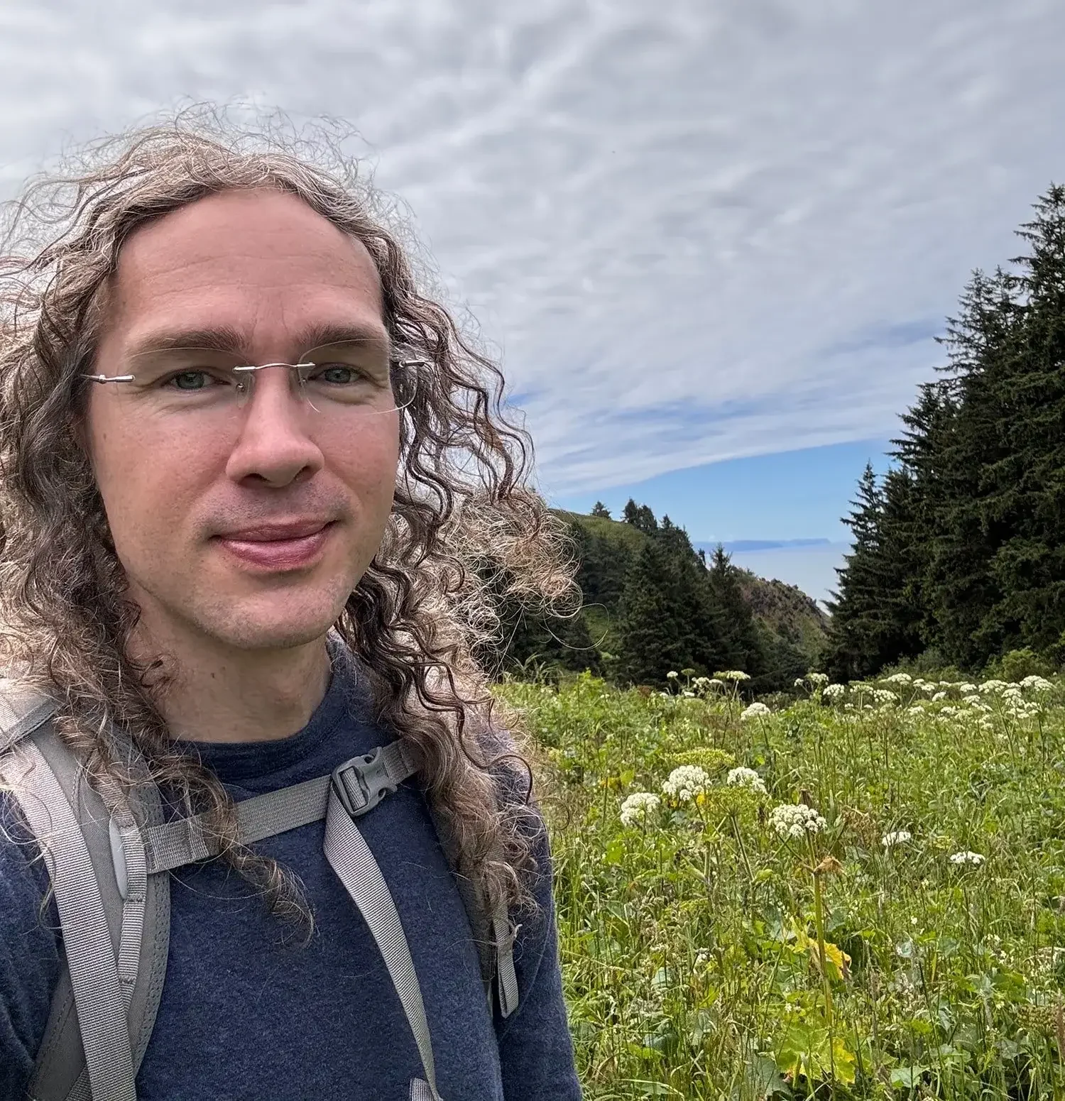 Jesse standing outdoors among green plants and white flowers, with a background of trees, hills, and a partly cloudy sky.