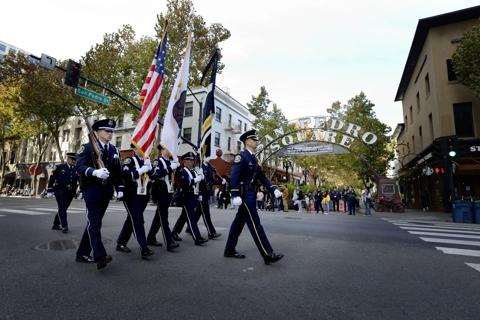 Police officers in uniform carrying flags in a parade on San Pedro Street in San Jose, CA with the San Pedro Square sign overhead and spectators watching on the sidewalk.