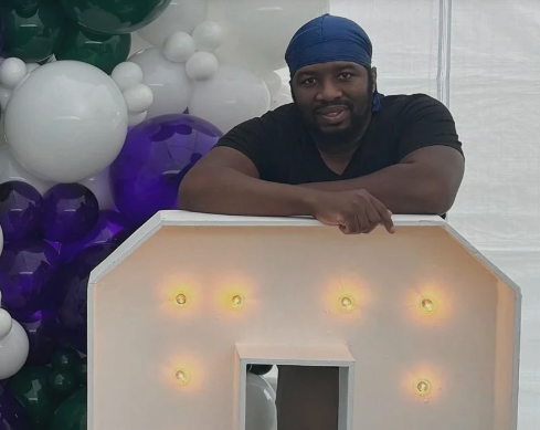 A man with a blue bandana resting his arms on a wooden structure with multiple small lights, with balloon decorations in white, purple, and green in the background.