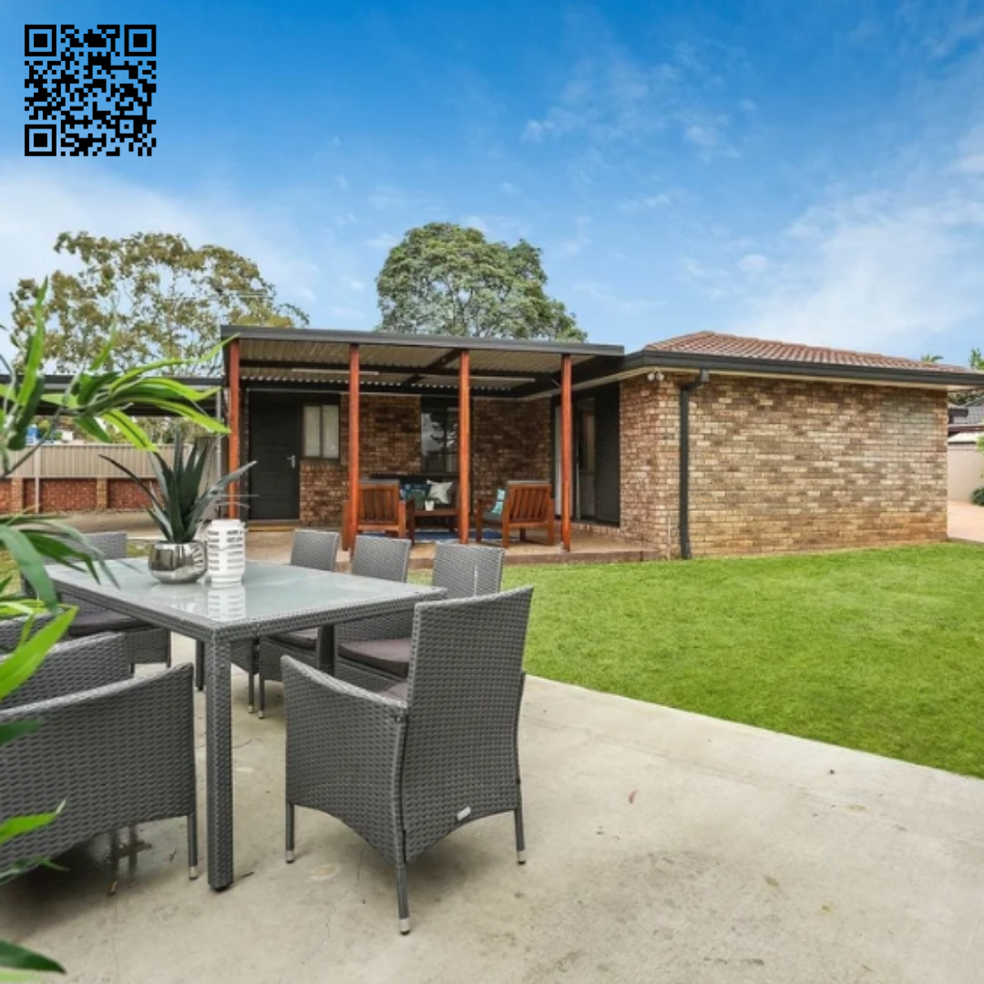 Backyard patio with outdoor dining table, chairs, and a covered seating area with wooden beams, grass lawn, and a brick house with a tiled roof under a blue sky.