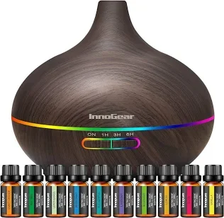 A dark-colored, round, ceramic essential oil diffuser with multiple colorful LED lights at the base and a variety of essential oils bottles lined up in front.