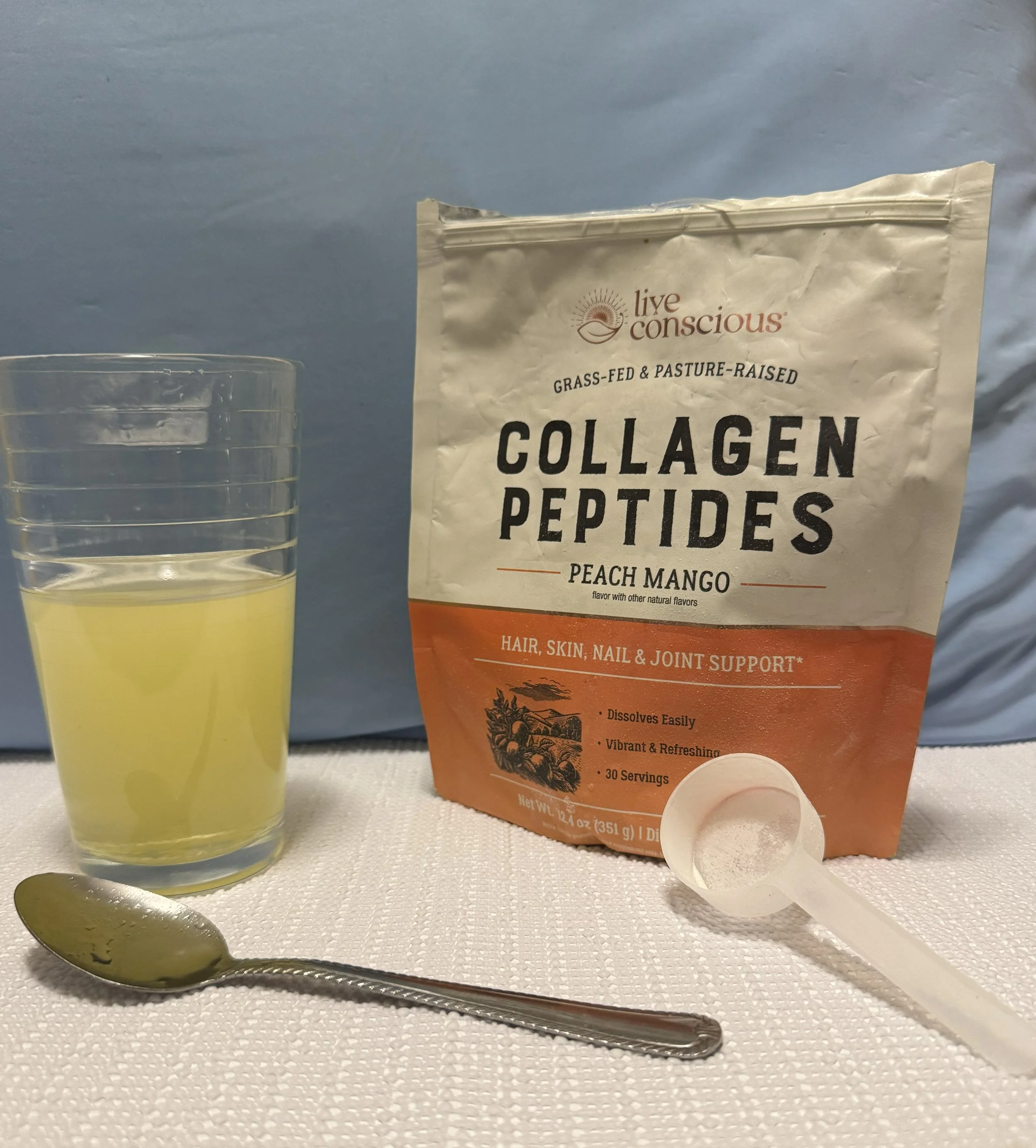 A packet of live conscious collagen peptides peach mango flavor alongside a glass of water, a silver spoon, and a white scoop on a white textured surface with a light blue background.