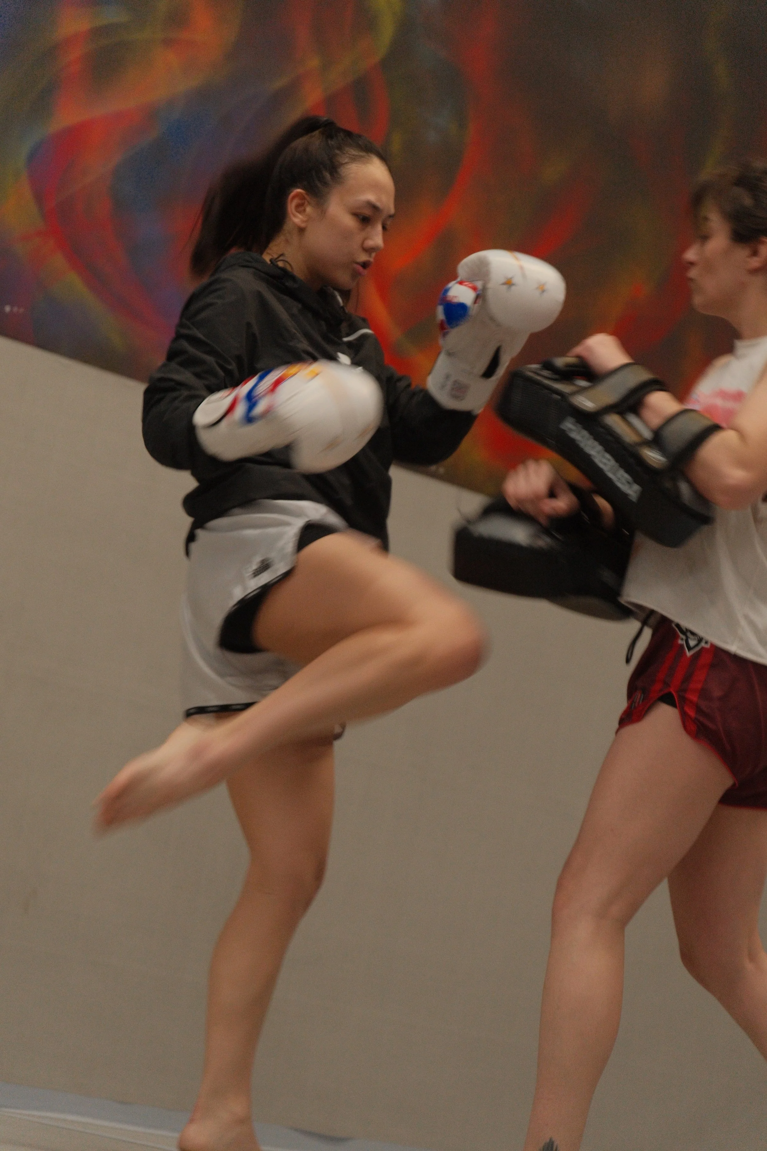 A woman practicing Muay Thai kicks while sparring with a trainer, wearing boxing gloves and athletic gear in a gym with abstract wall art in the background.