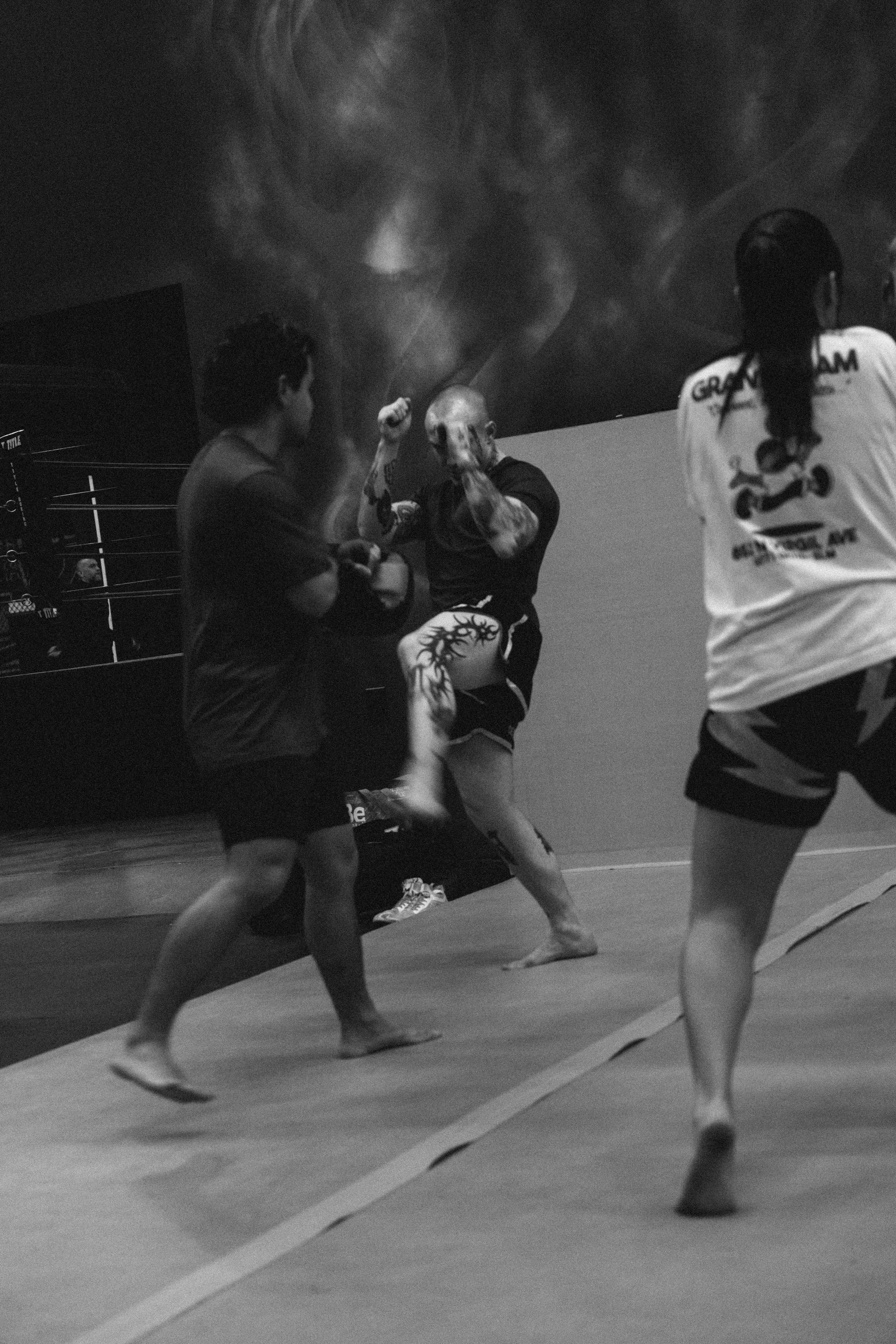 Three women practicing Muay Thai in a gym, with one sparring partner and a large projected image of a boxer on the wall behind them.