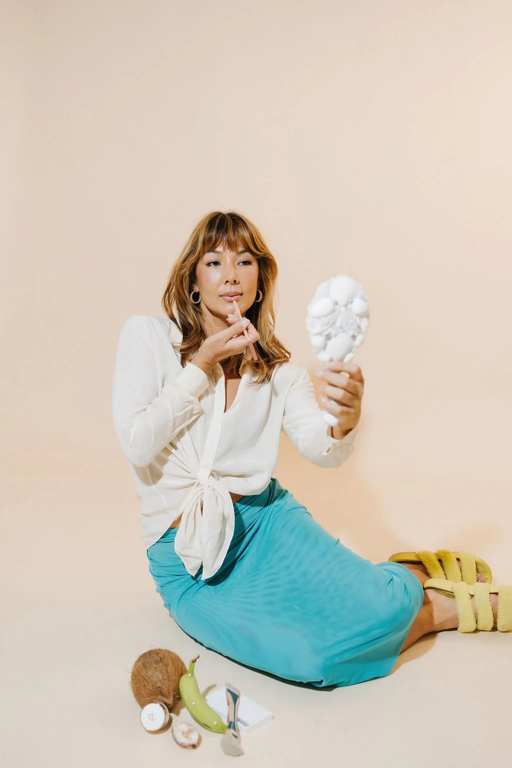 Woman in white blouse and blue skirt, sitting on the floor, looking at a mirror and applying lip gloss, with tropical fruits including a banana, coconut, and skincare products nearby.