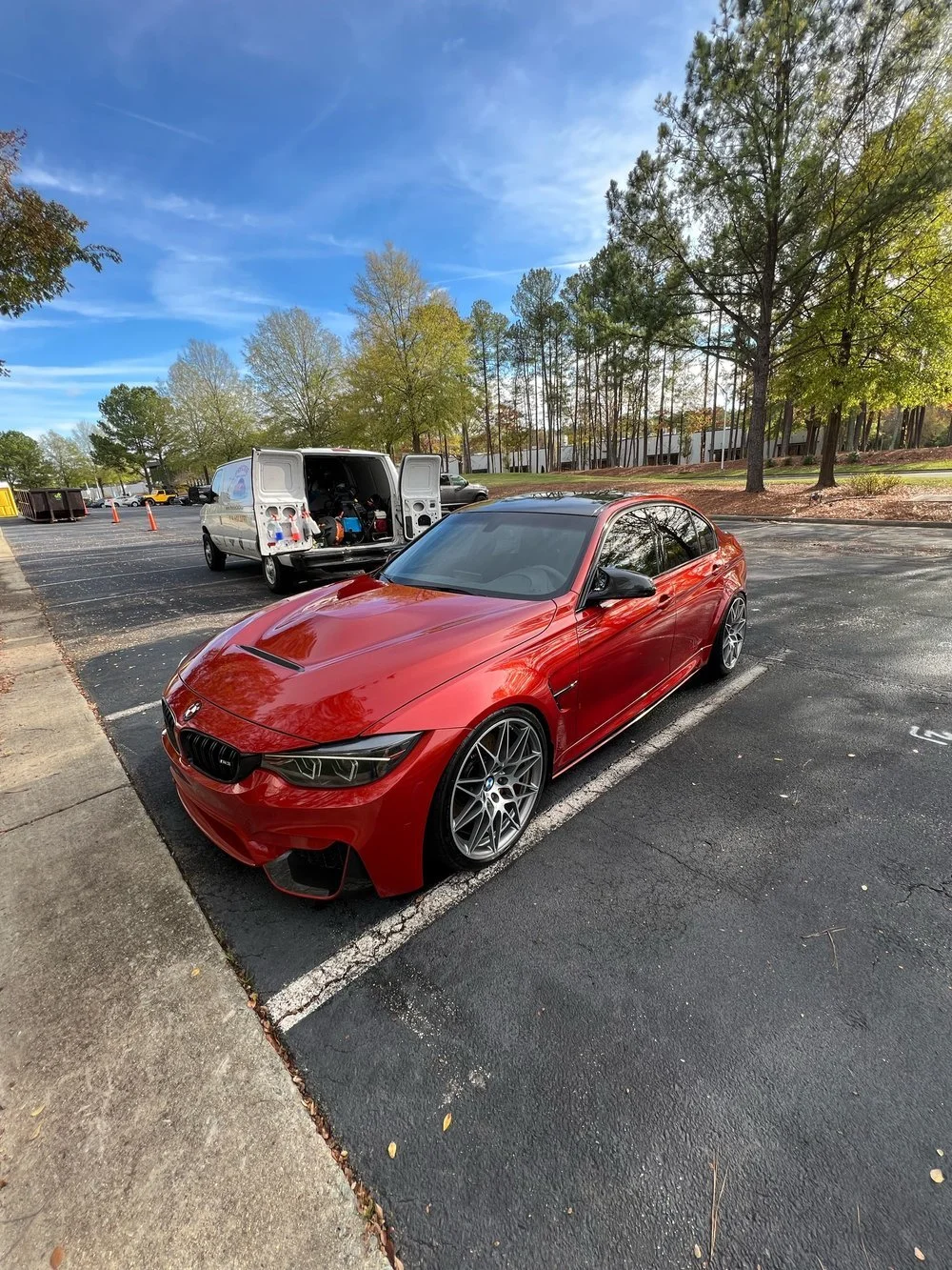 Red BMW parked in a parking lot with trees and a white van in the background, on a clear day.