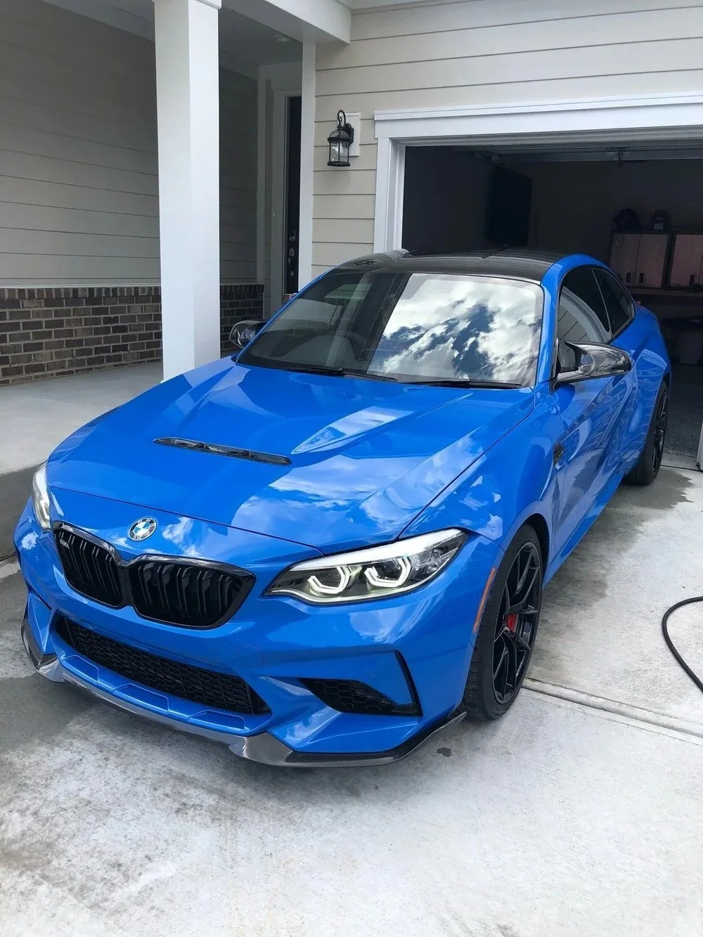 A blue BMW M4 parked in a driveway outside a house with its garage door open, reflecting the sky on the windshield.