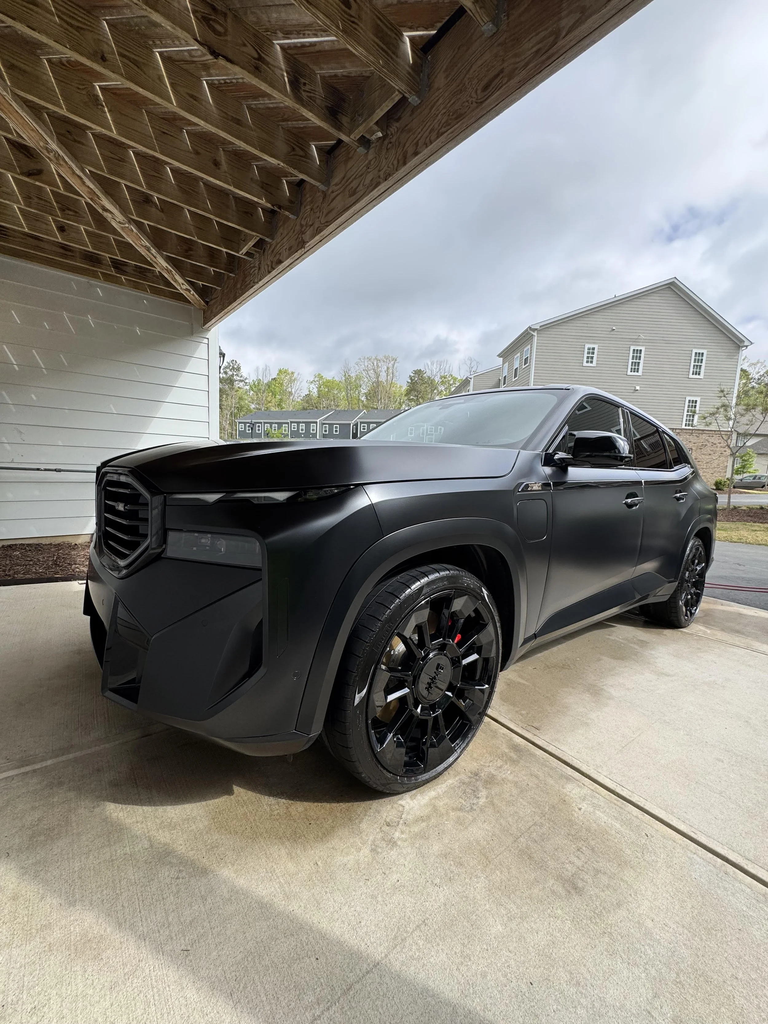 A black futuristic SUV parked under a wooden carport with residential buildings and trees in the background.