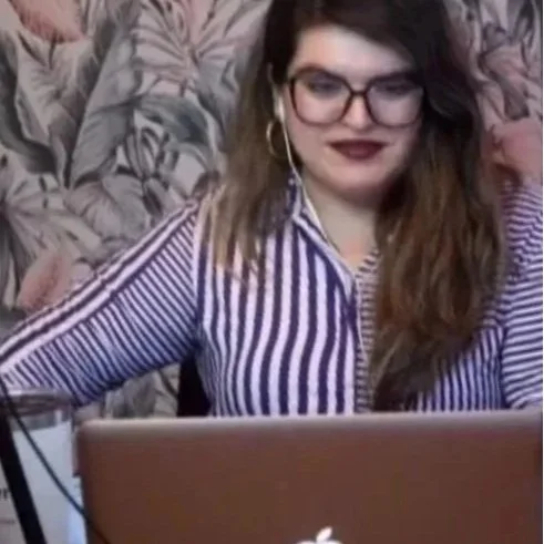 Mujer con gafas y orejera, sonriendo y usando una blusa a rayas, trabajando frente a una computadora portátil.