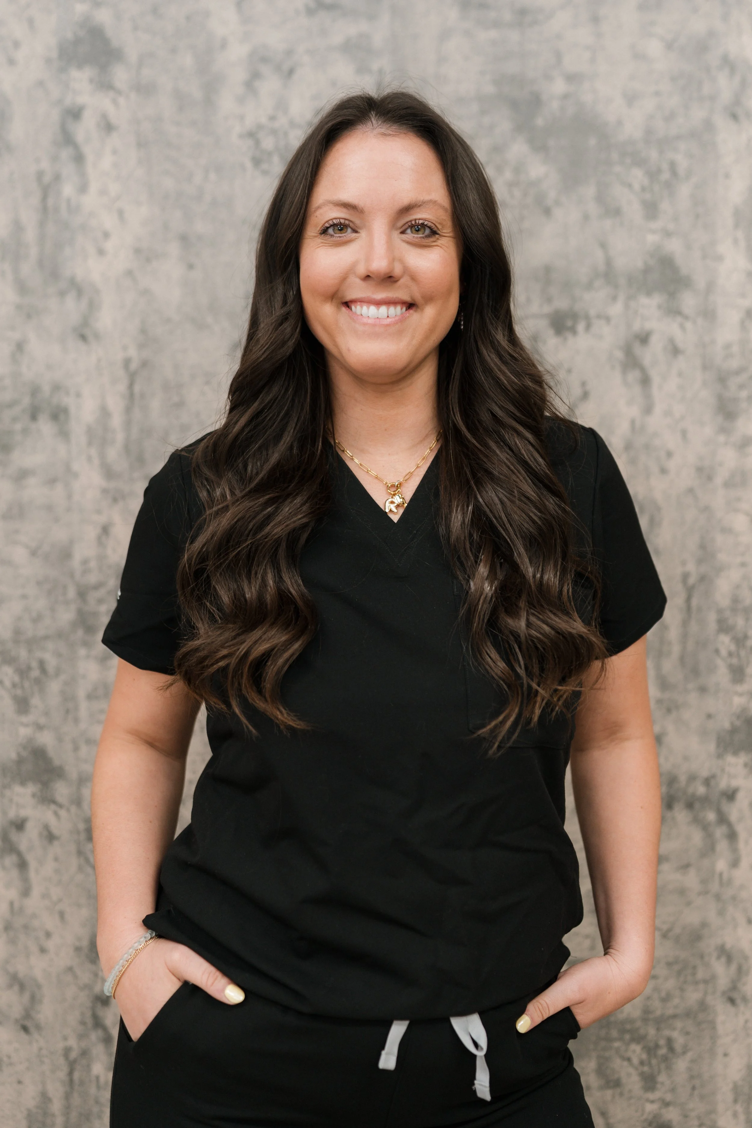 Smiling person with long brown hair, wearing a black top, against a light-colored wall background.