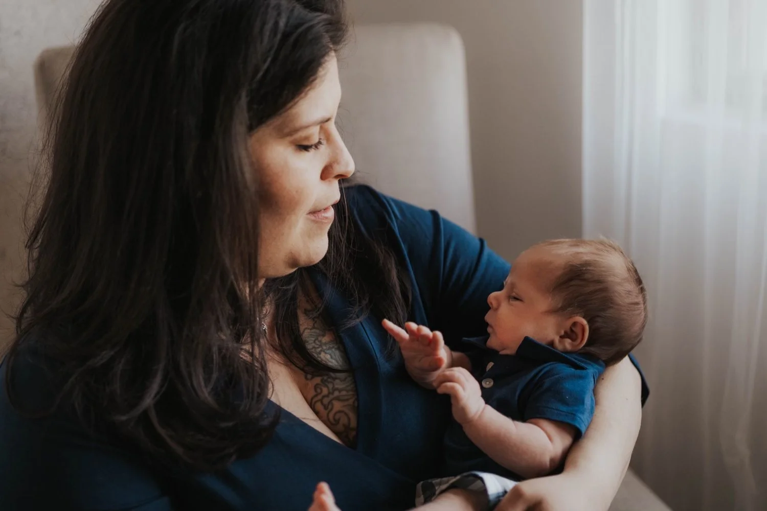 A female client of Stephanie Celis, a lactation consultant in Gilbert Arizona, holding a baby in her arms, both looking at each other near a window with curtains.