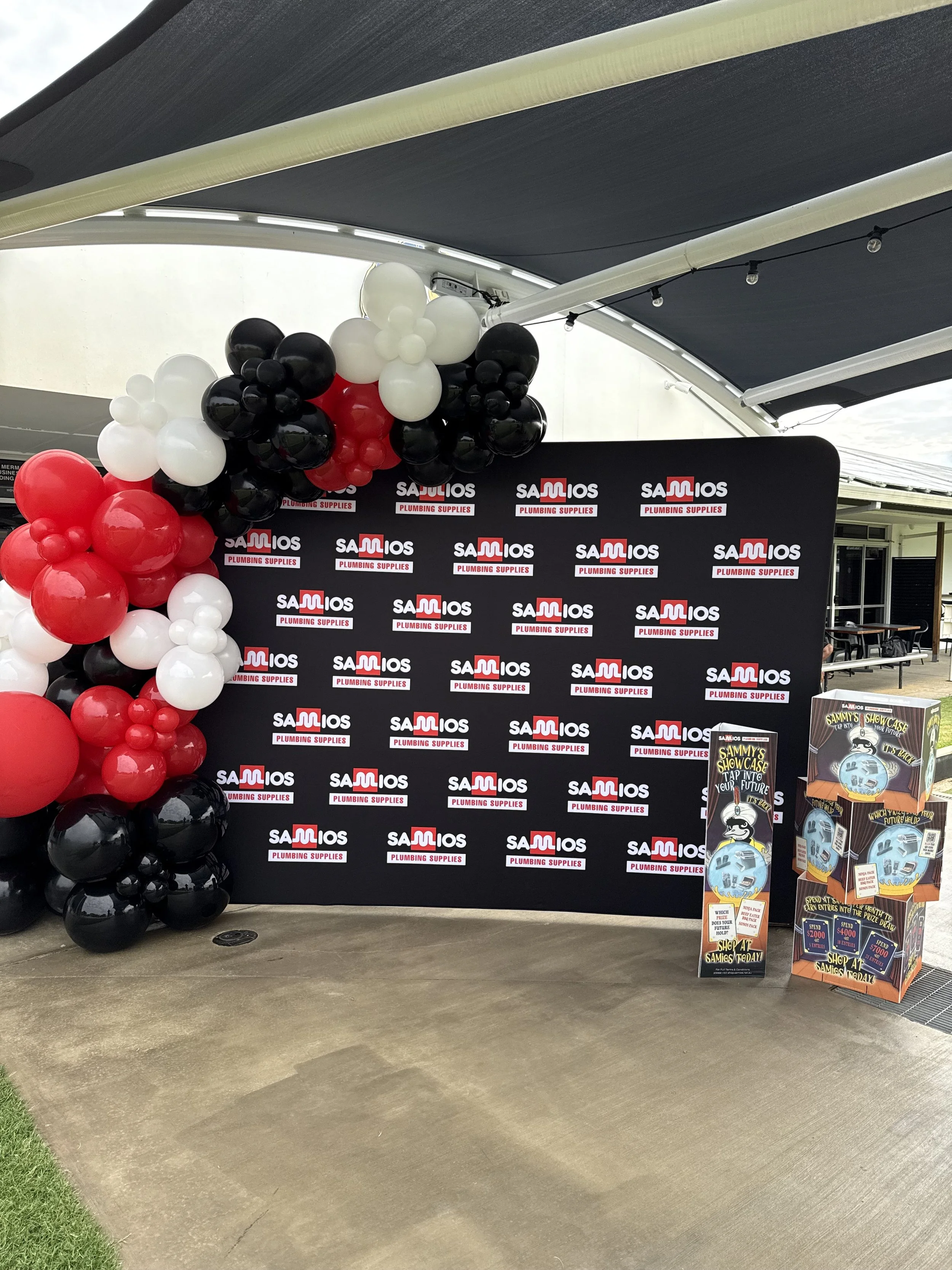 Black, white, and red balloons arranged near a black backdrop with 'Saulios Plumbing Supplies' logos, under a shaded area.