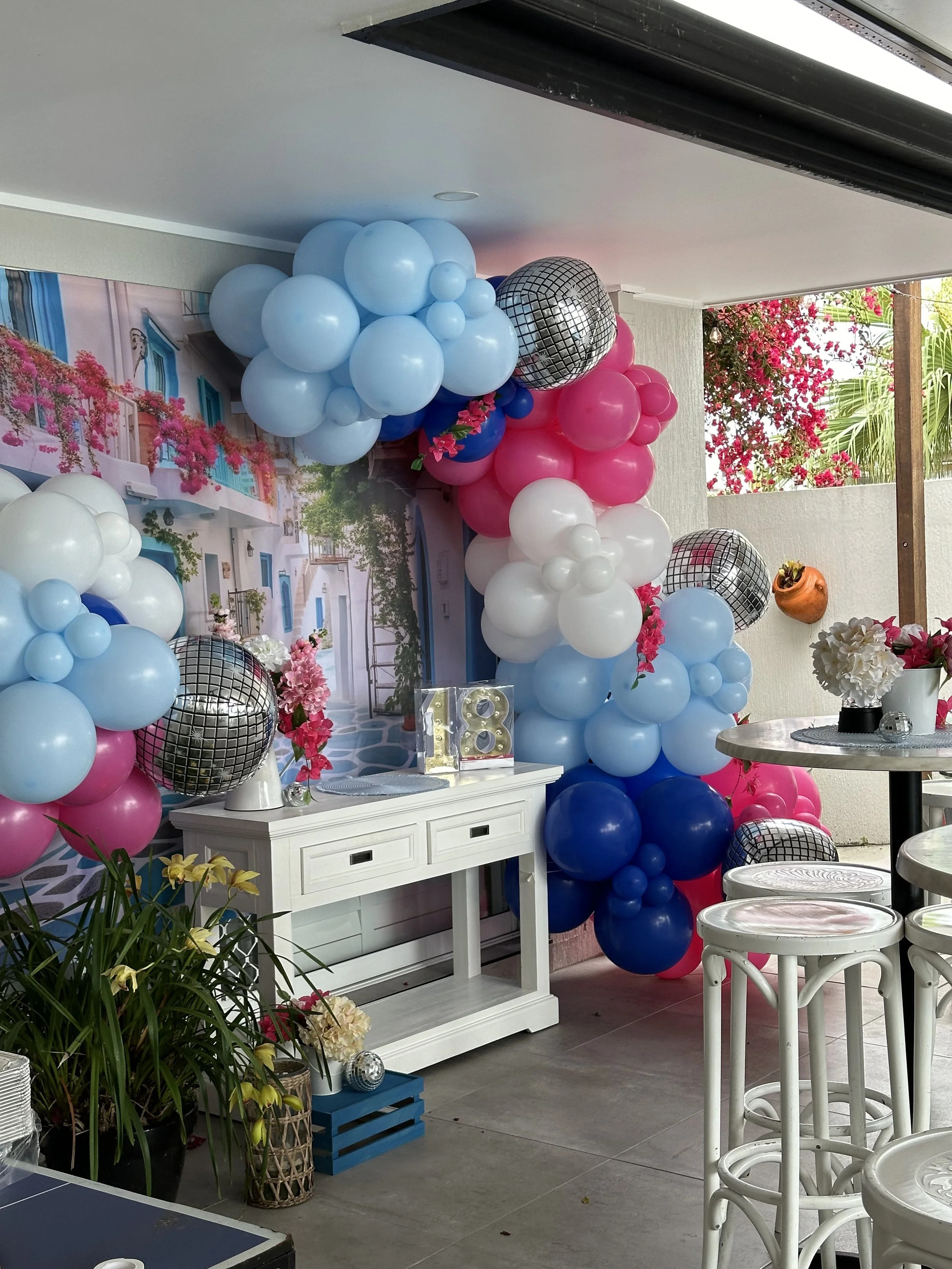 Decorative 'Mamma Mia' themed party setup with pink, navy, light blue, white, and black disco ball balloons, a backdrop of a coastal scene with pink flowers.