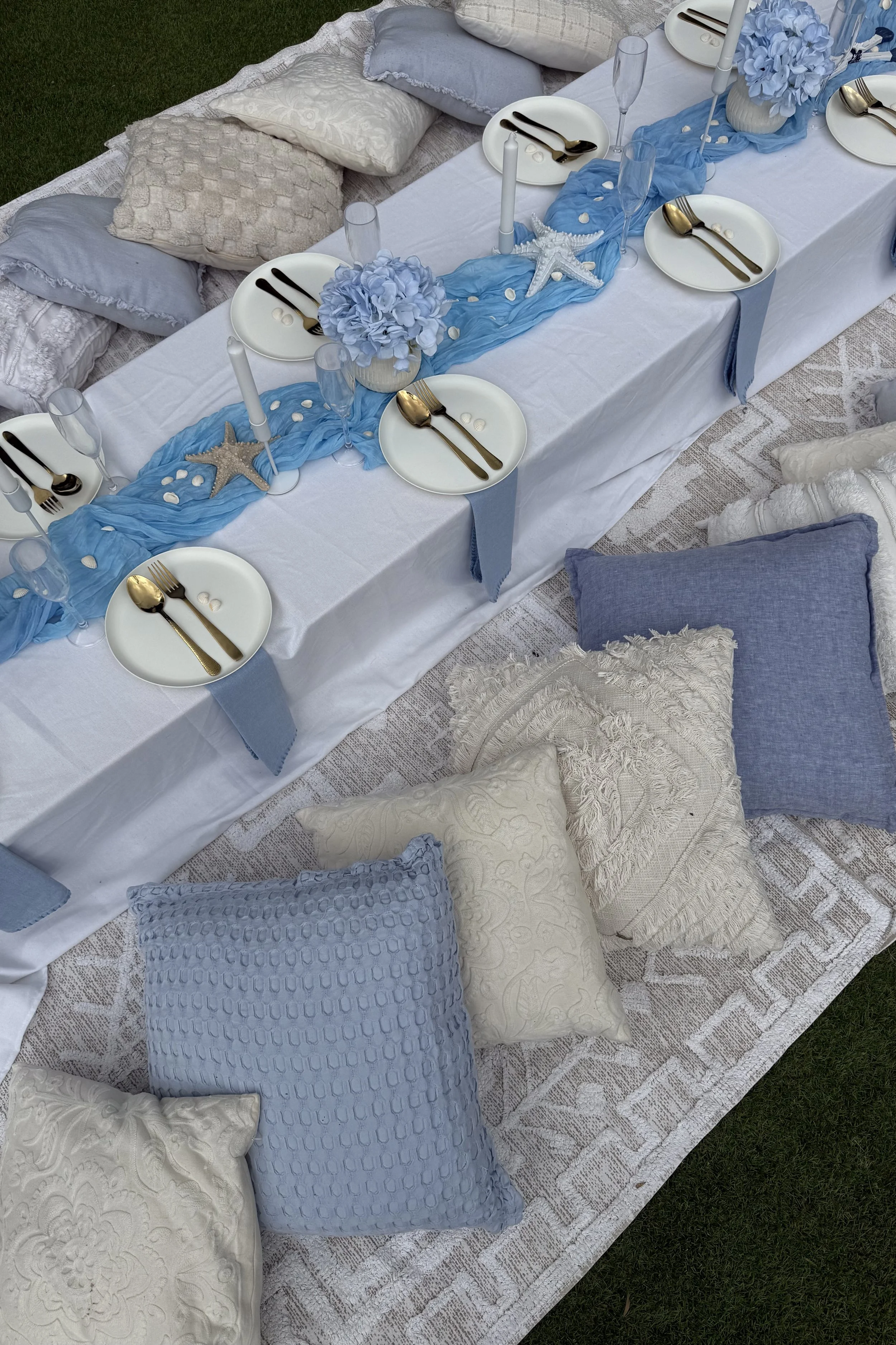 A table set for a meal with blue and white decor, including plates with gold utensils, blue table runner, flower arrangements, and sitting on a rug surrounded by cushions.