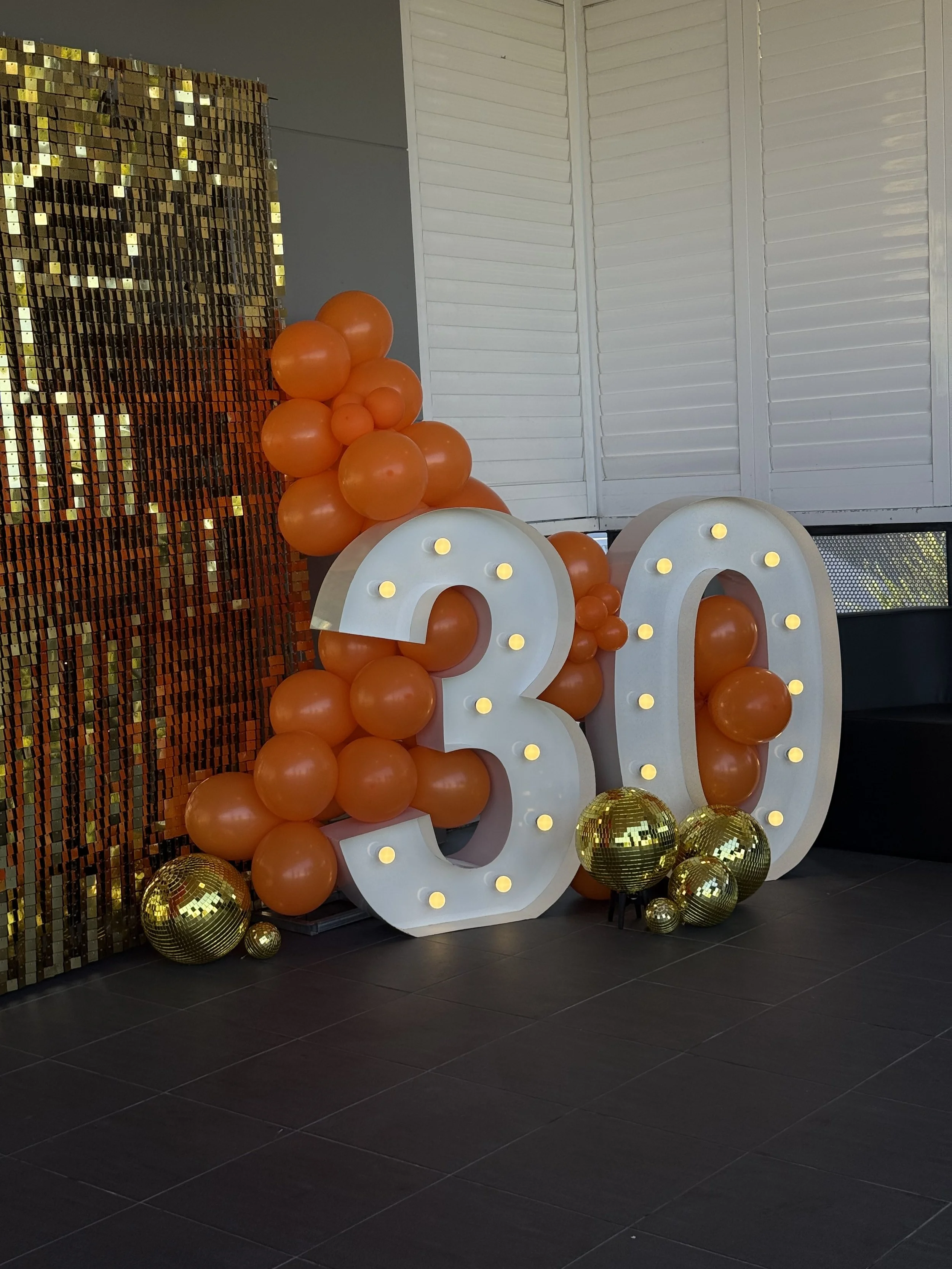 Large illuminated number 30 with orange balloons and gold decorations behind and around, indicating a 30th celebration or anniversary.