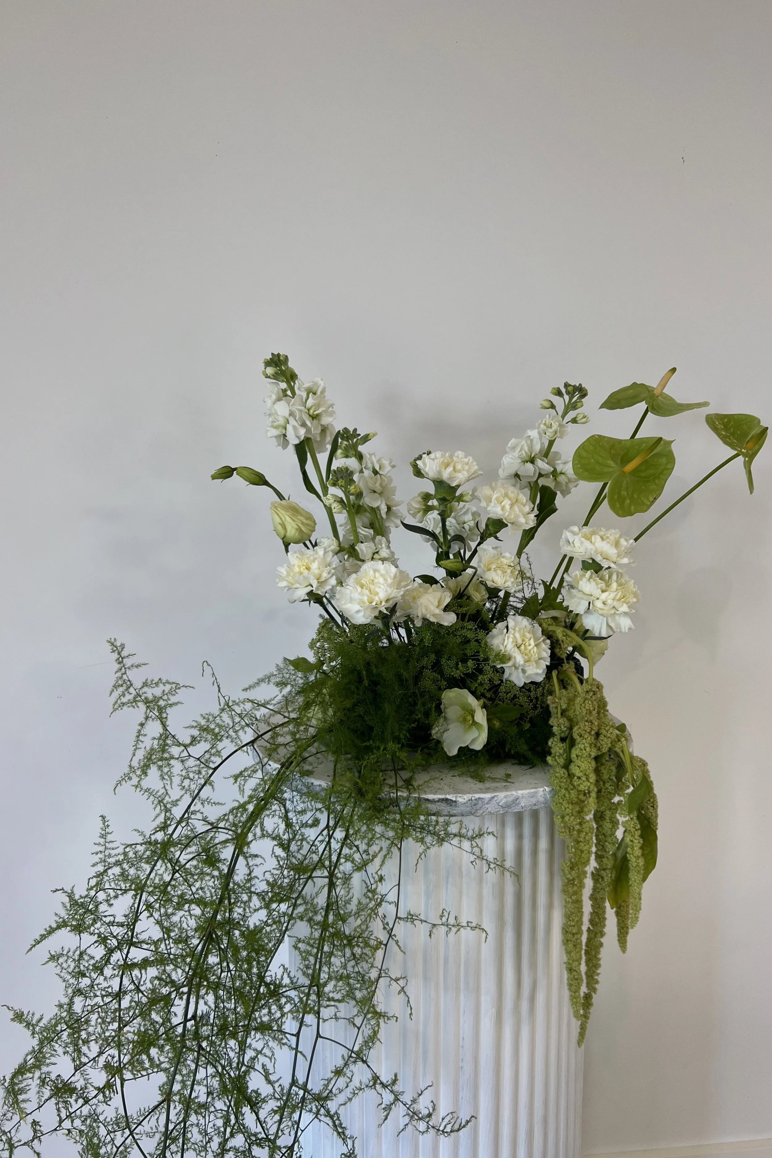 White floral arrangement with various flowers and green foliage on a white textured pedestal against a plain light-colored wall.