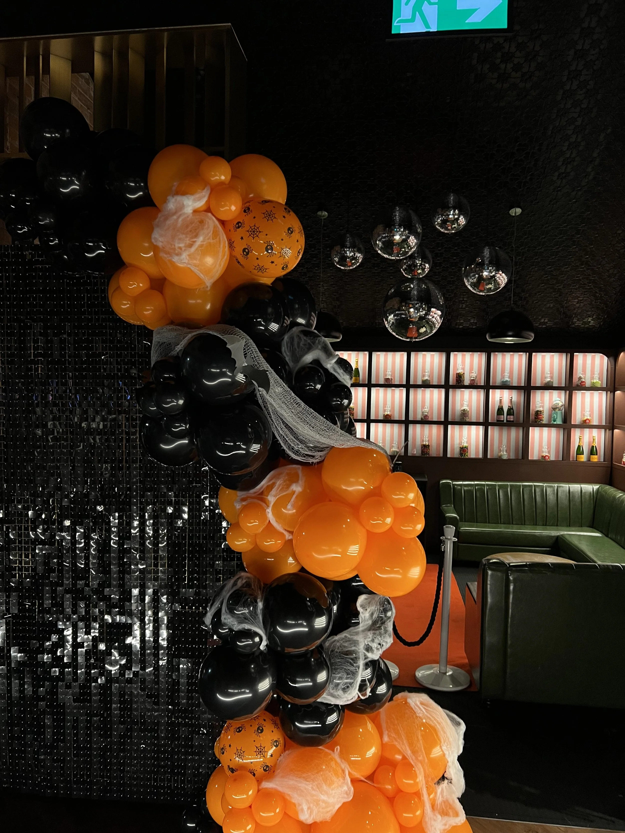 Halloween-themed balloon decoration with black and orange balloons, decorated with cobwebs, in a modern indoor space with a black wall, mirrored hanging balls.