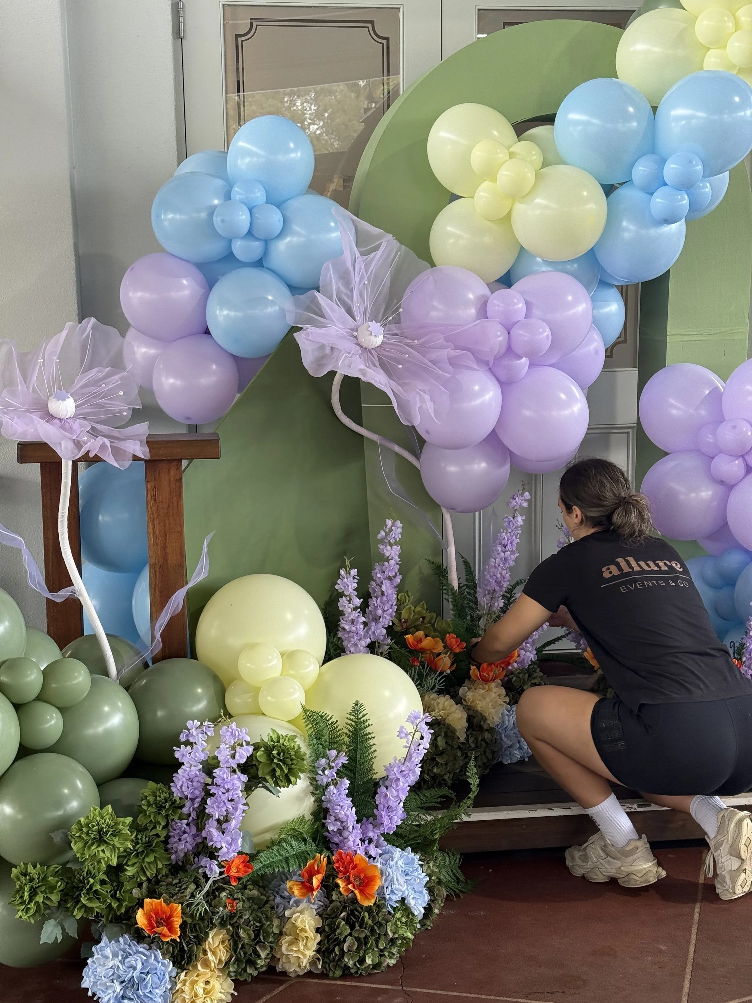 A colorful balloon garland and floral arrangment. The display features purple, green, blue, and yellow balloons, some arranged in clusters, along with a variety of artificial flowers including lavender, orange, blue, and cream colors.