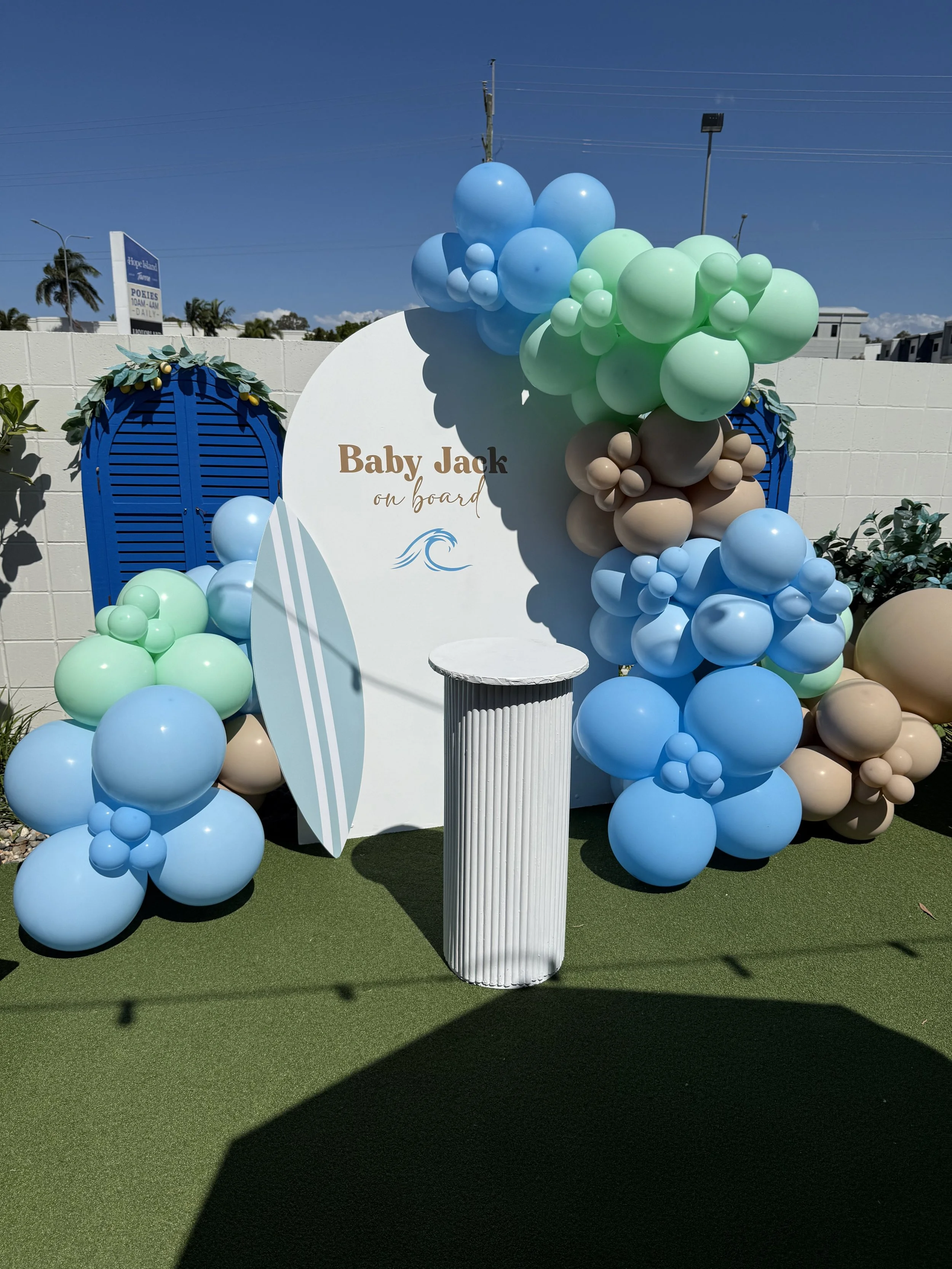 Decorative ballon garland on a timber arch with a surfboard theme for a boy baby shower using blues, greens and beige balloon colours, featuring a ripple plinth for a cake display.