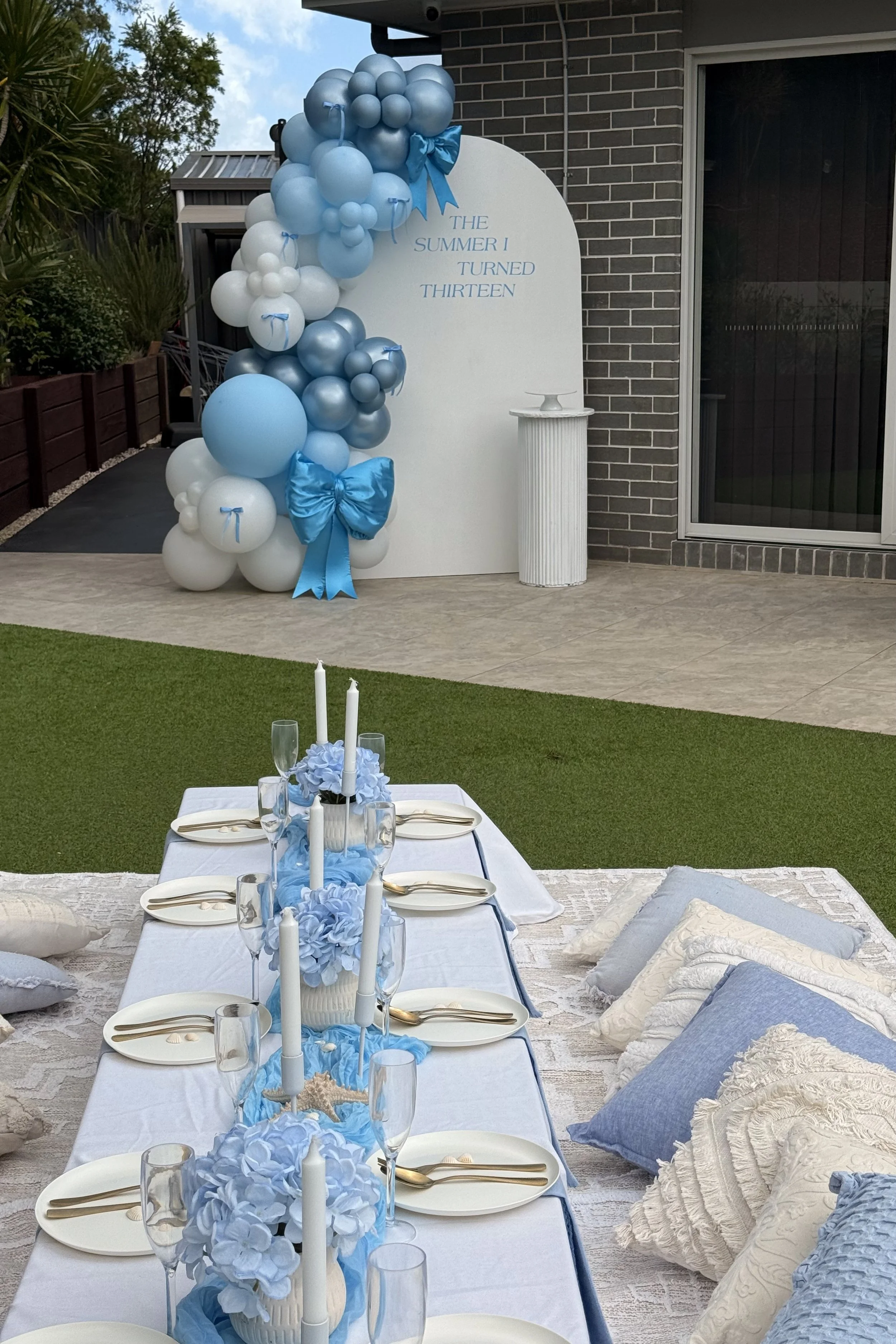 Outdoor celebration setup with a table decorated in blue and white, featuring candles, hydrangeas, and gold cutlery, adjacent to a white backdrop with blue balloons and a sign reading 'The Summer I Turned Thirteen'.