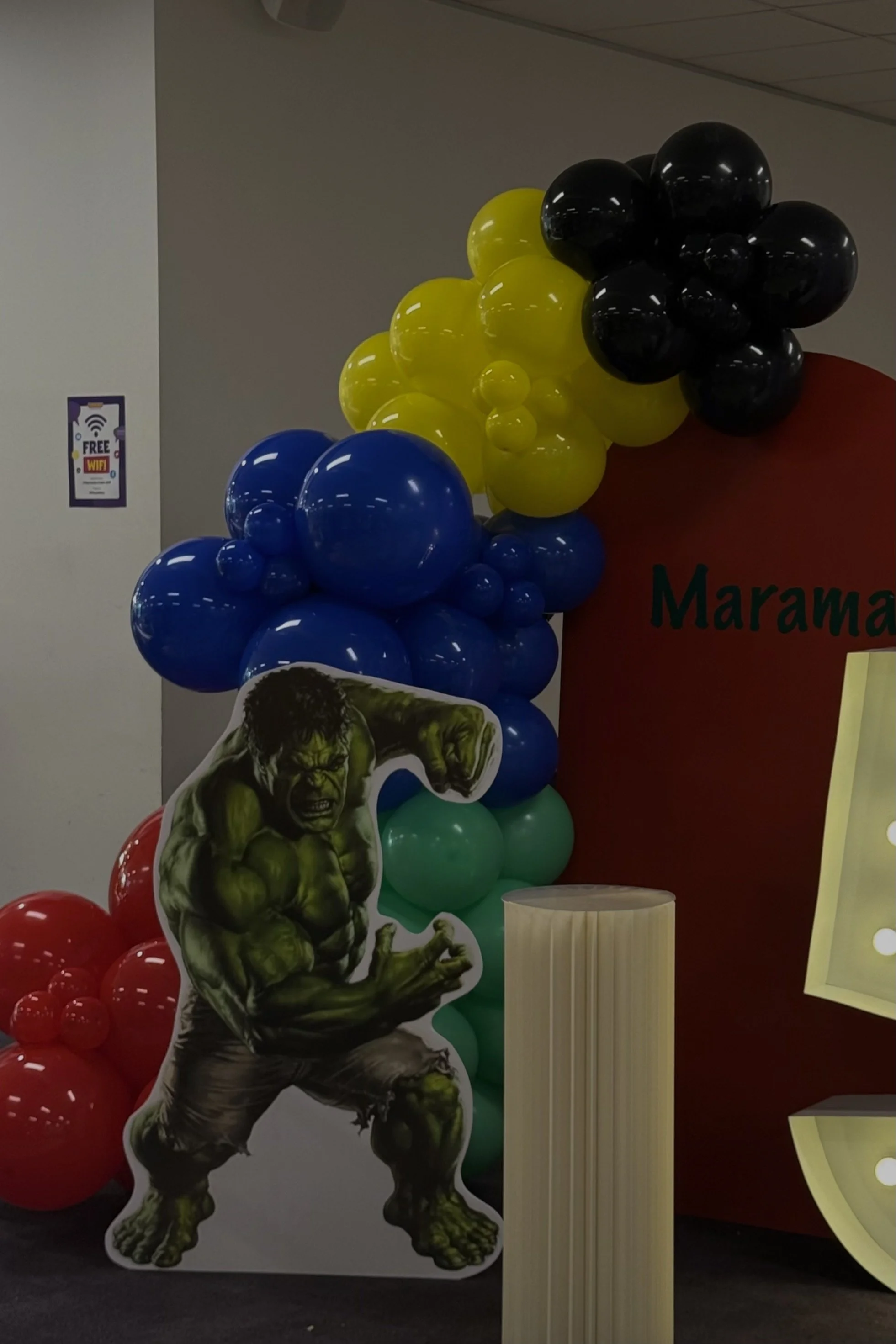 Colorful balloon decorations including red, green, blue, yellow, and black balloons arranged in clusters with a large cutout of the Hulk showing a fierce expression and pose.