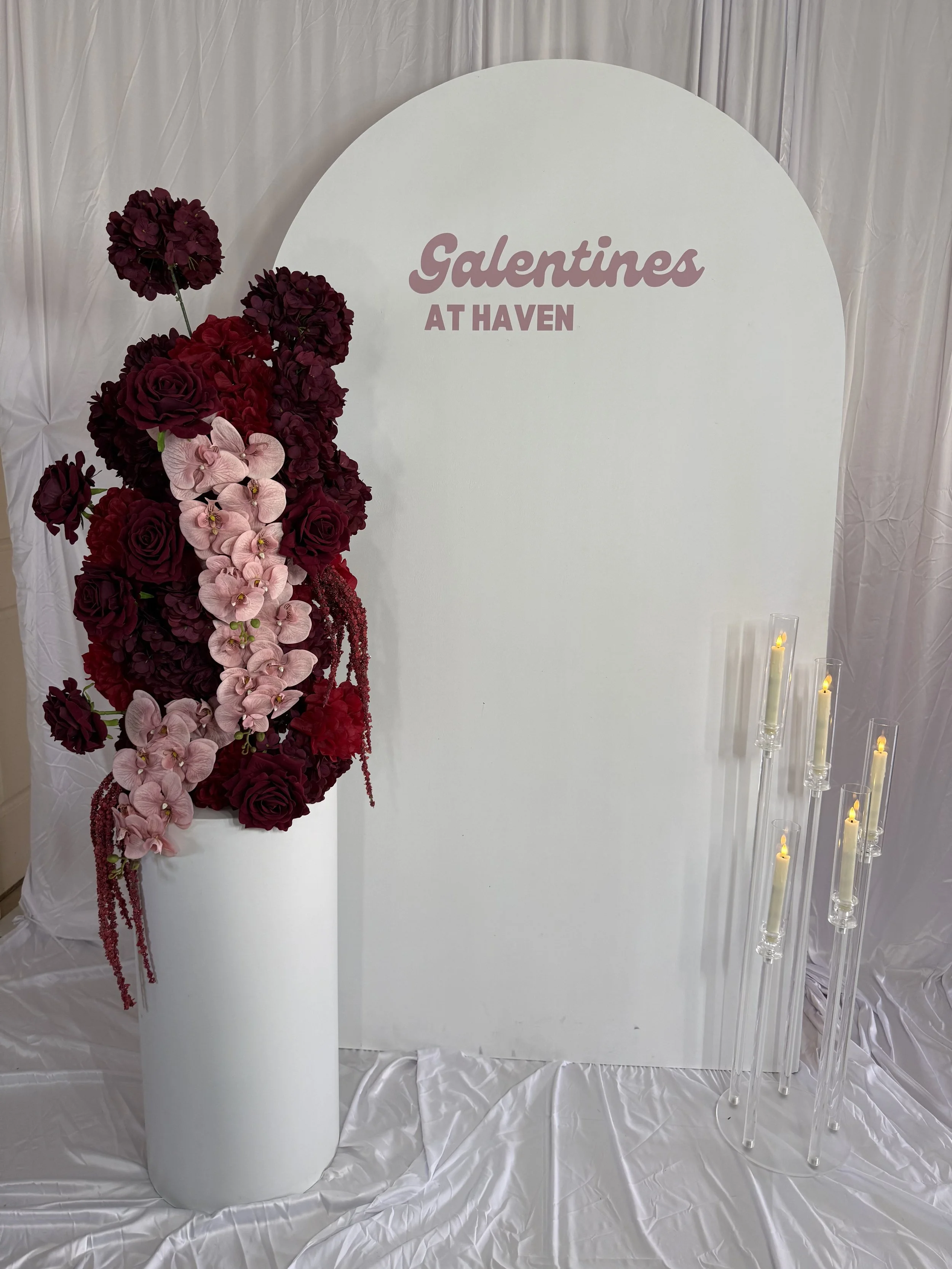 A white backdrop with the words 'Galentines at Haven' in pink and purple. In front, there's a tall white vase with a large arrangement of burgundy roses, pink orchids, and maroon flowers. To the right, there are tall clear candle holders with flicker