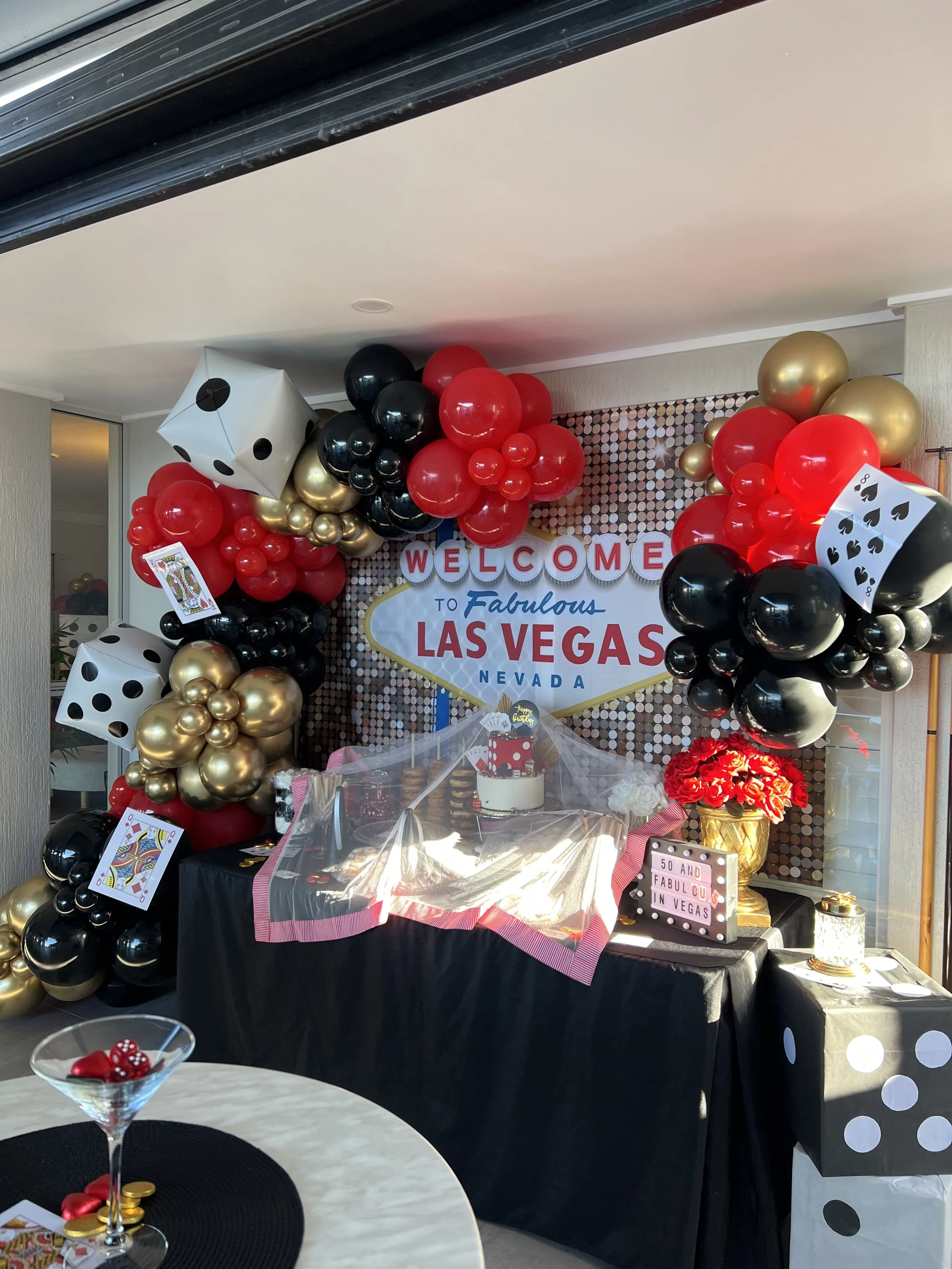 Decorative Poker themed backdrop with a balloon garland in black, red, and gold colours, featuring large dice and playing cards, with a sign saying 'Welcome to Fabulous Las Vegas Nevada'. 