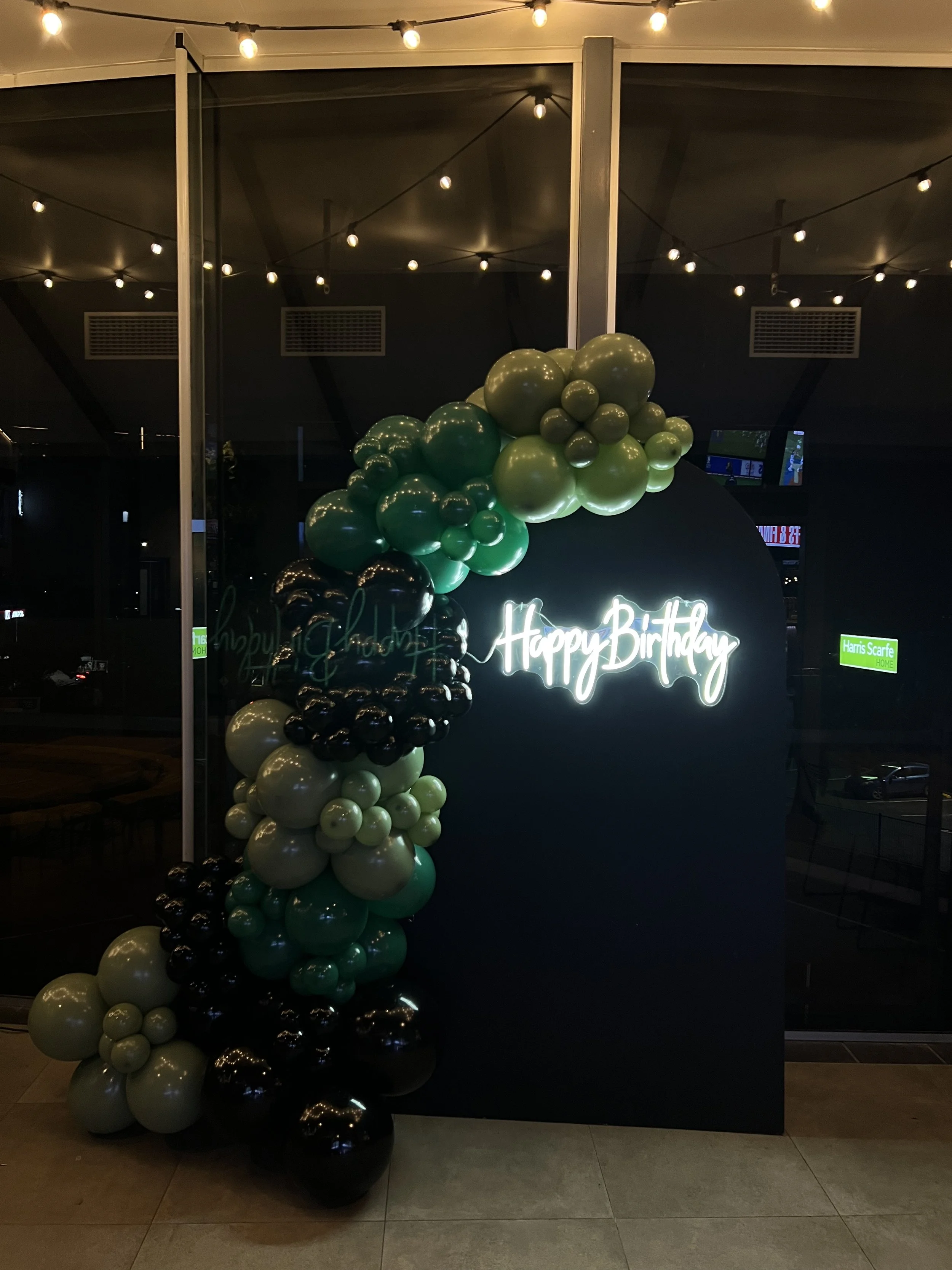A balloon decoration with black, dark green, light green, and beige balloons arranged along a black backdrop with a neon "Happy Birthday" sign.