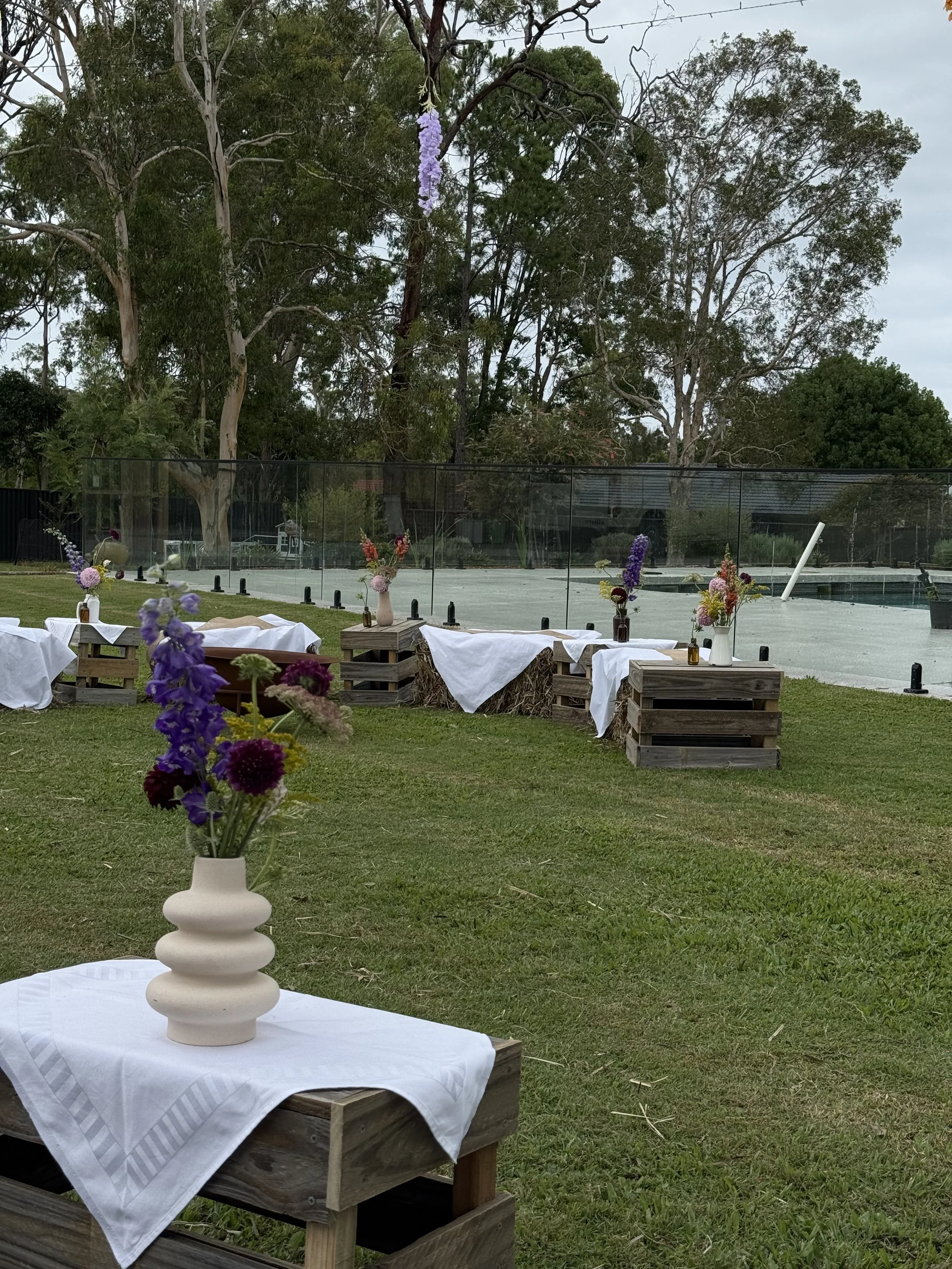 Outdoor event setup with tables and flower arrangements on wooden pallets with white tablecloths, near a tennis court and surrounded by trees.