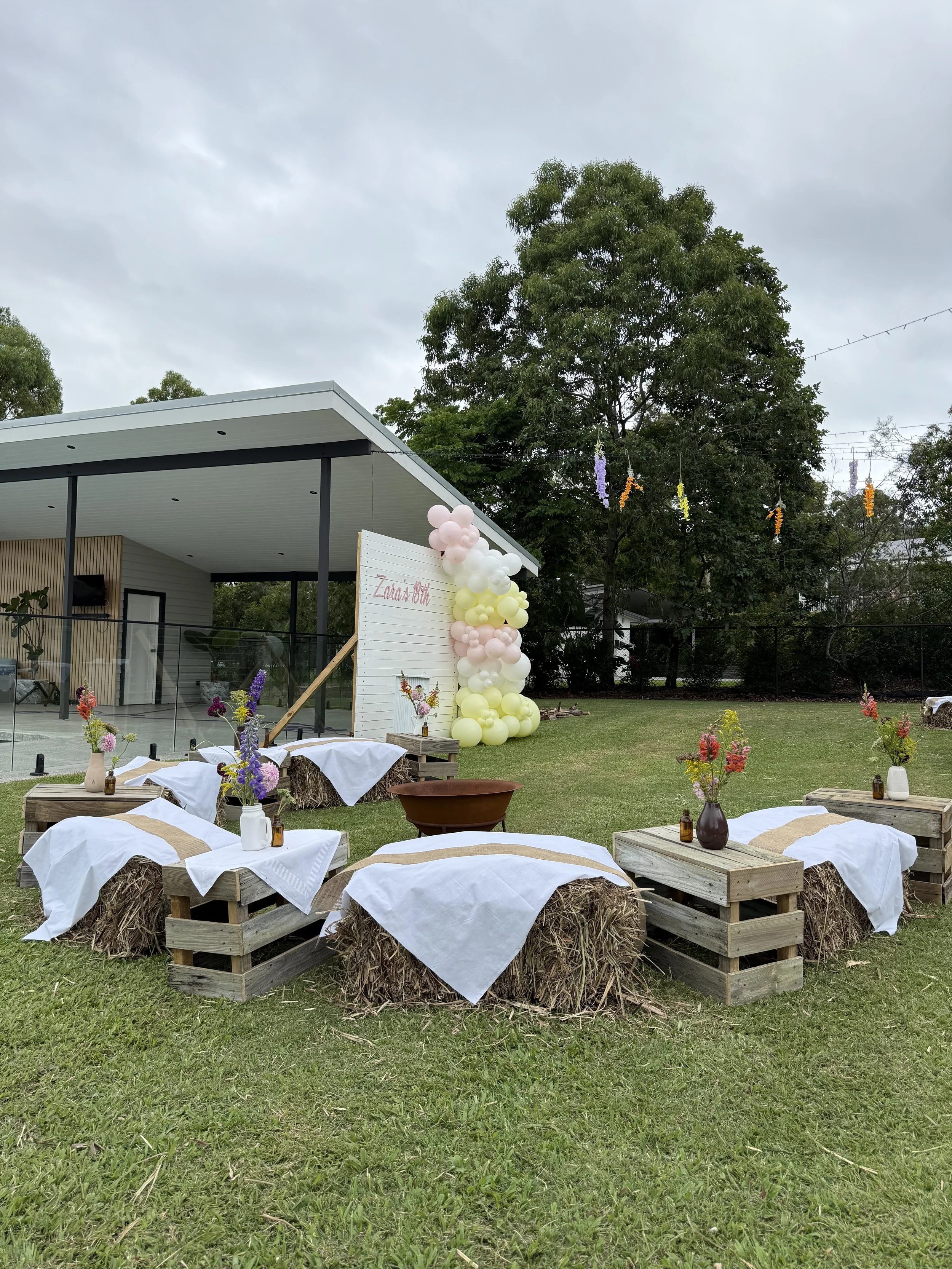Outdoor setting with hay bales covered with white cloths, decorated with vases of colorful flowers, arranged around a grassy yard. In the background, there is a modern building with a small deck, and a decorative arch made of pink, white, and yellow 