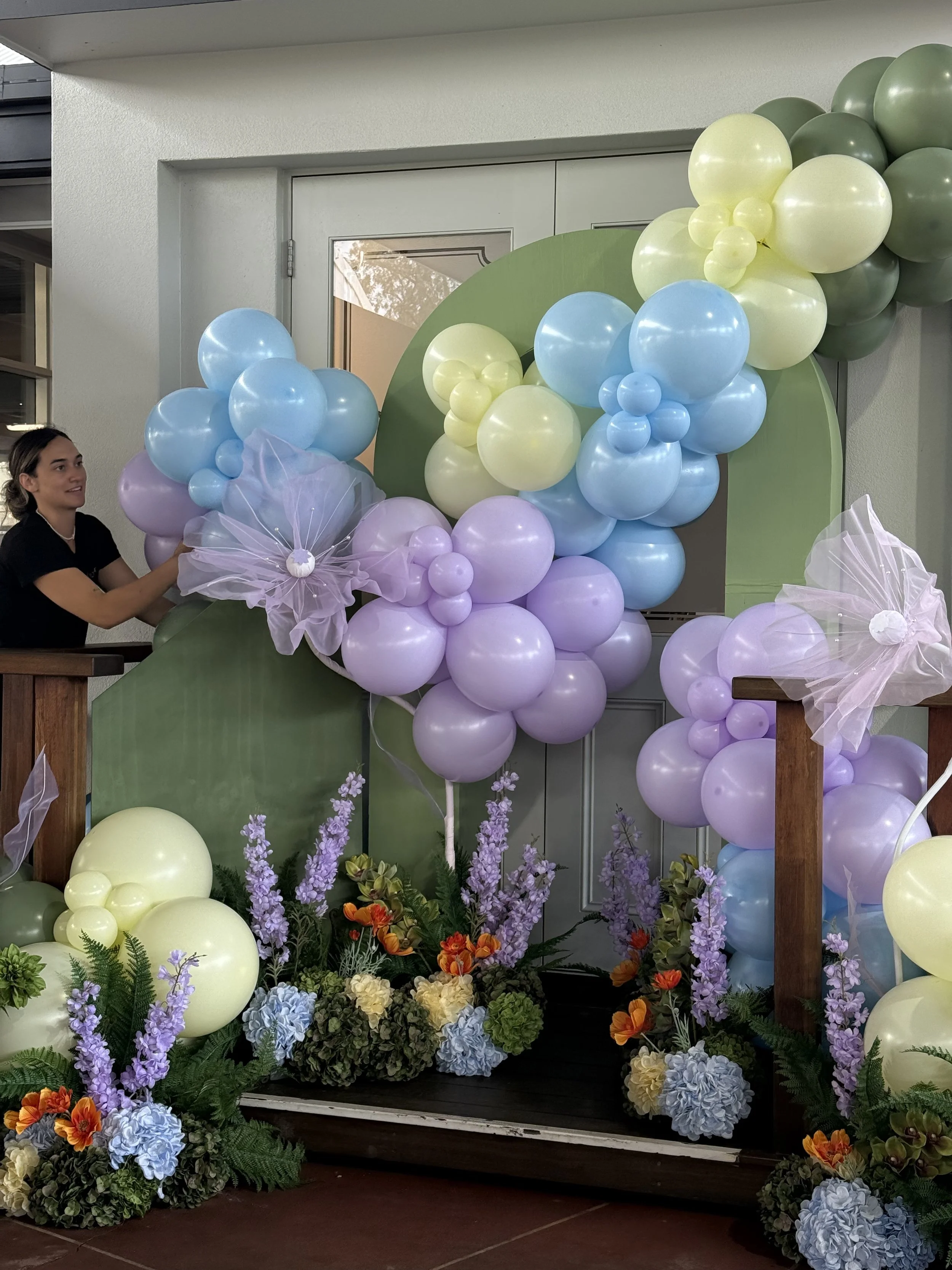 Decorative balloon arch with pastel blue, purple, yellow, and green balloons, accented by giant purple tulle flowers , and floral arrangements including lavender, orange, yellow, and blue flowers with greenery.