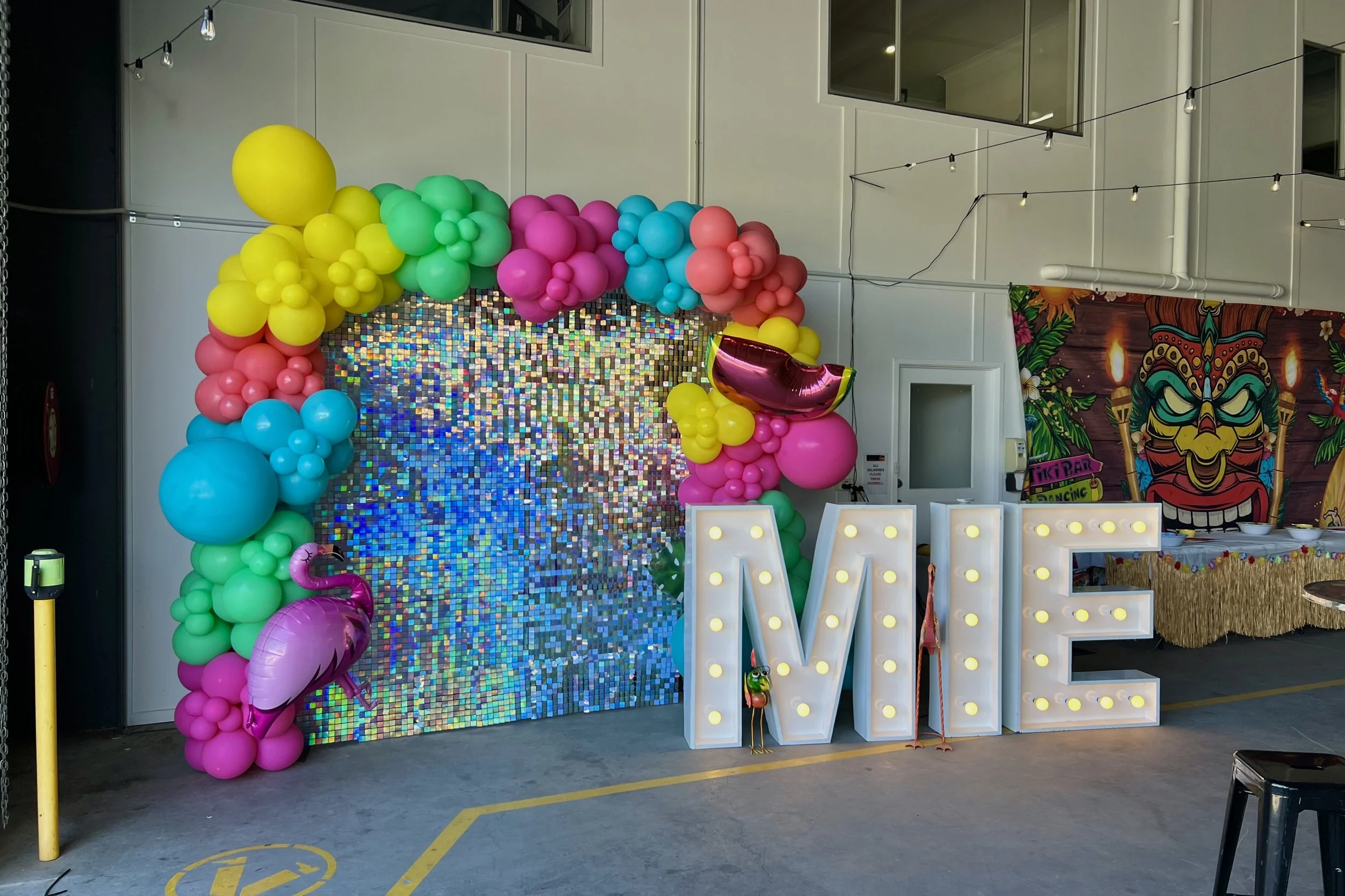 Colourful balloon garland on a shimmer wall backdrop with large lightup letters.