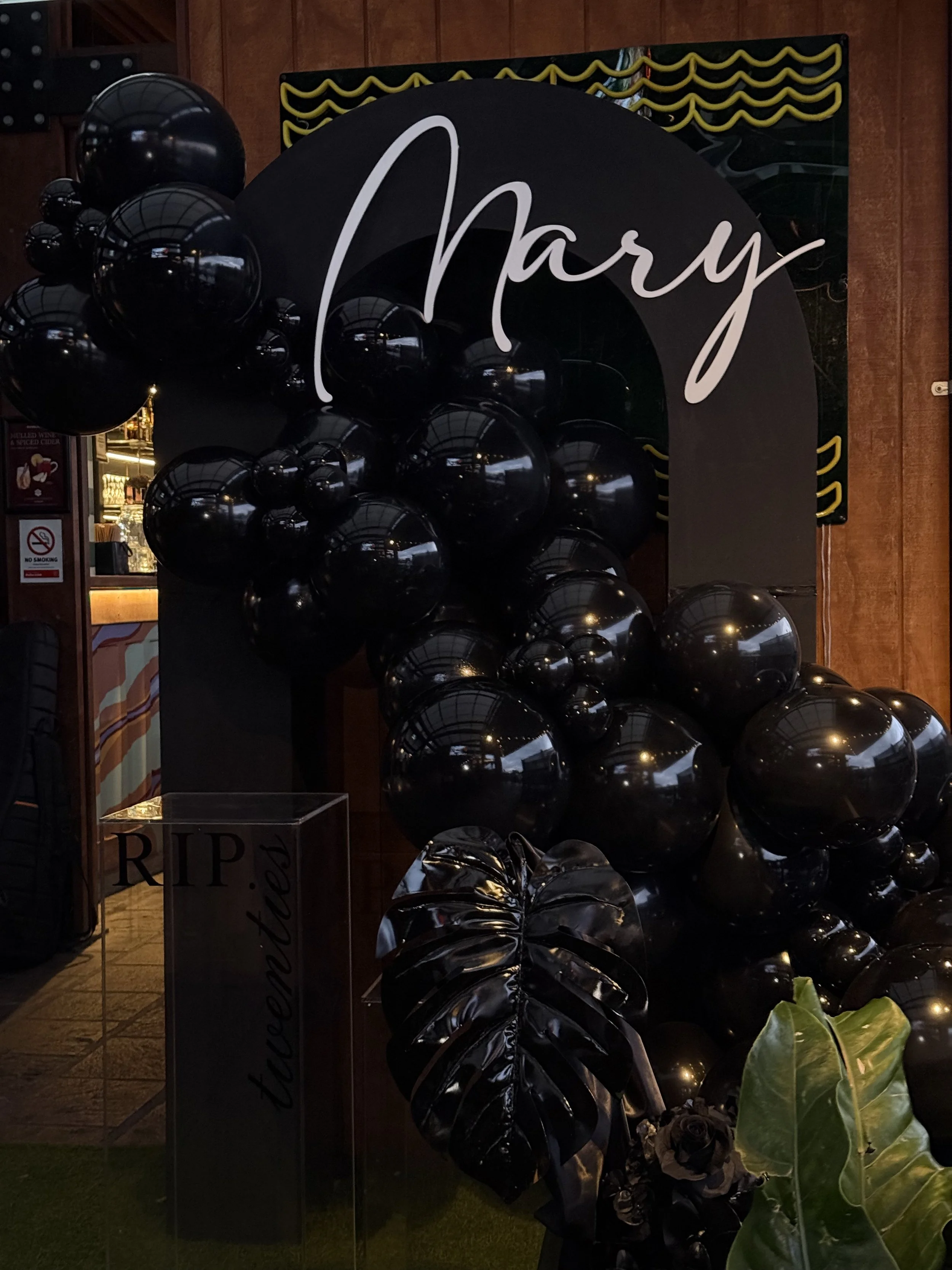 Black hollow arch backdrop with a shiny black ballon garland, acrylic name sign with greennery and a black ripple plinth.