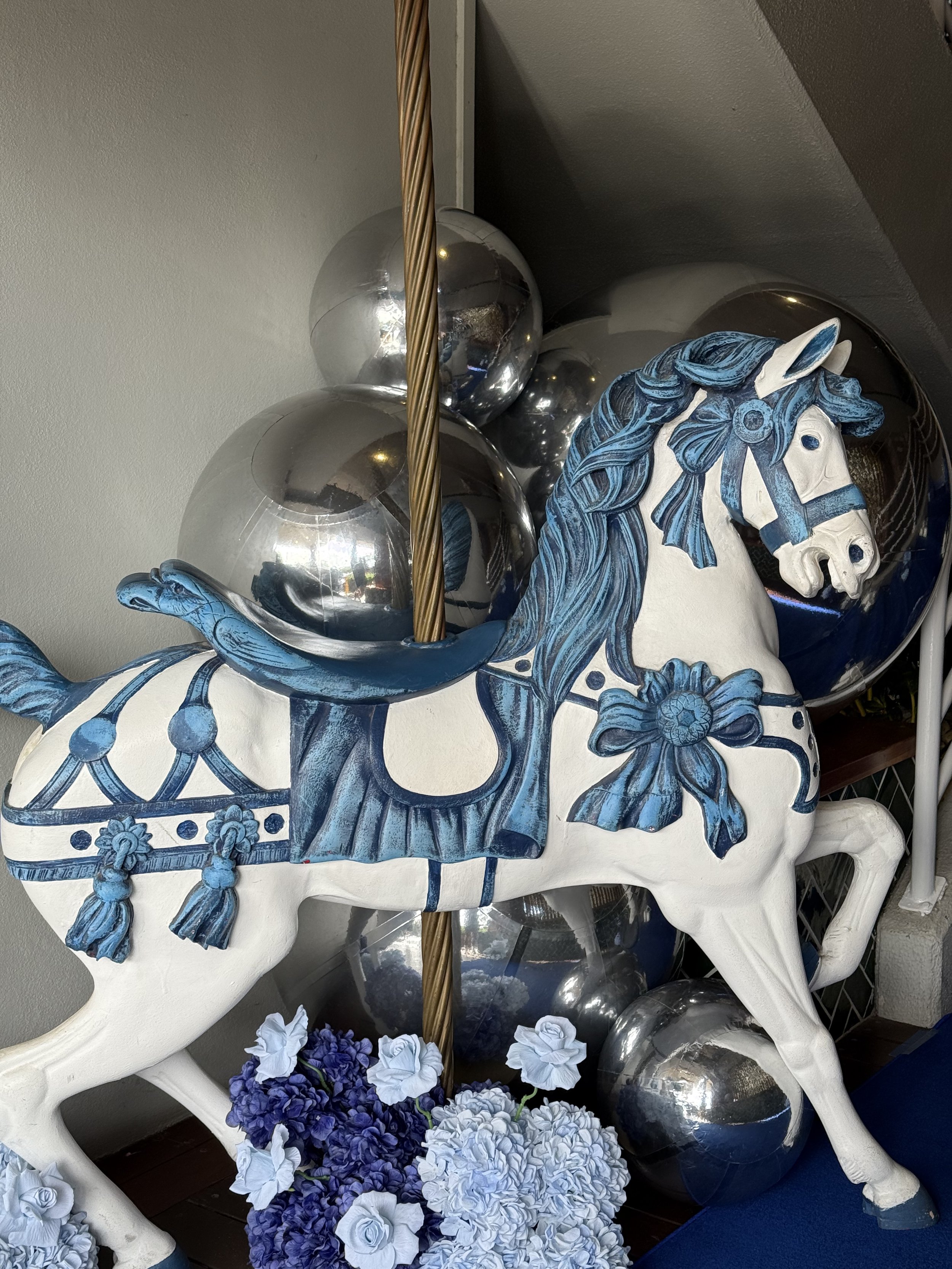 Decorative carousel horse sculpture painted white with blue accents, surrounded by big shiny silver balls and navy and light blue artificial flowers