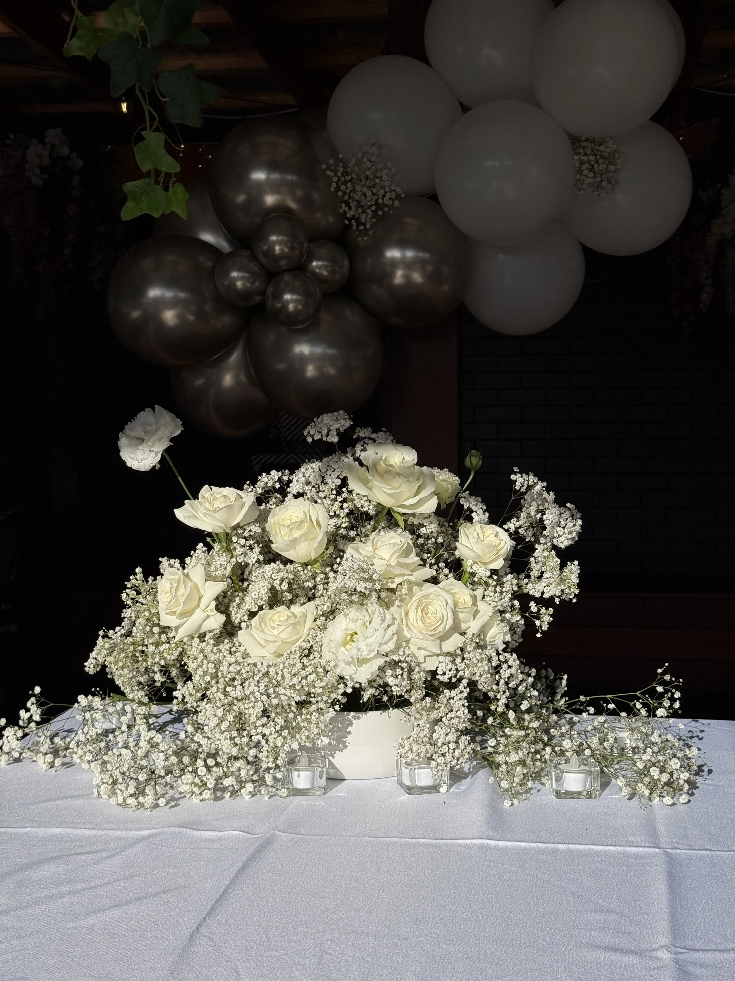 A floral arrangement of white roses and baby's breath on a white table, with white and silver balloons in the background.
